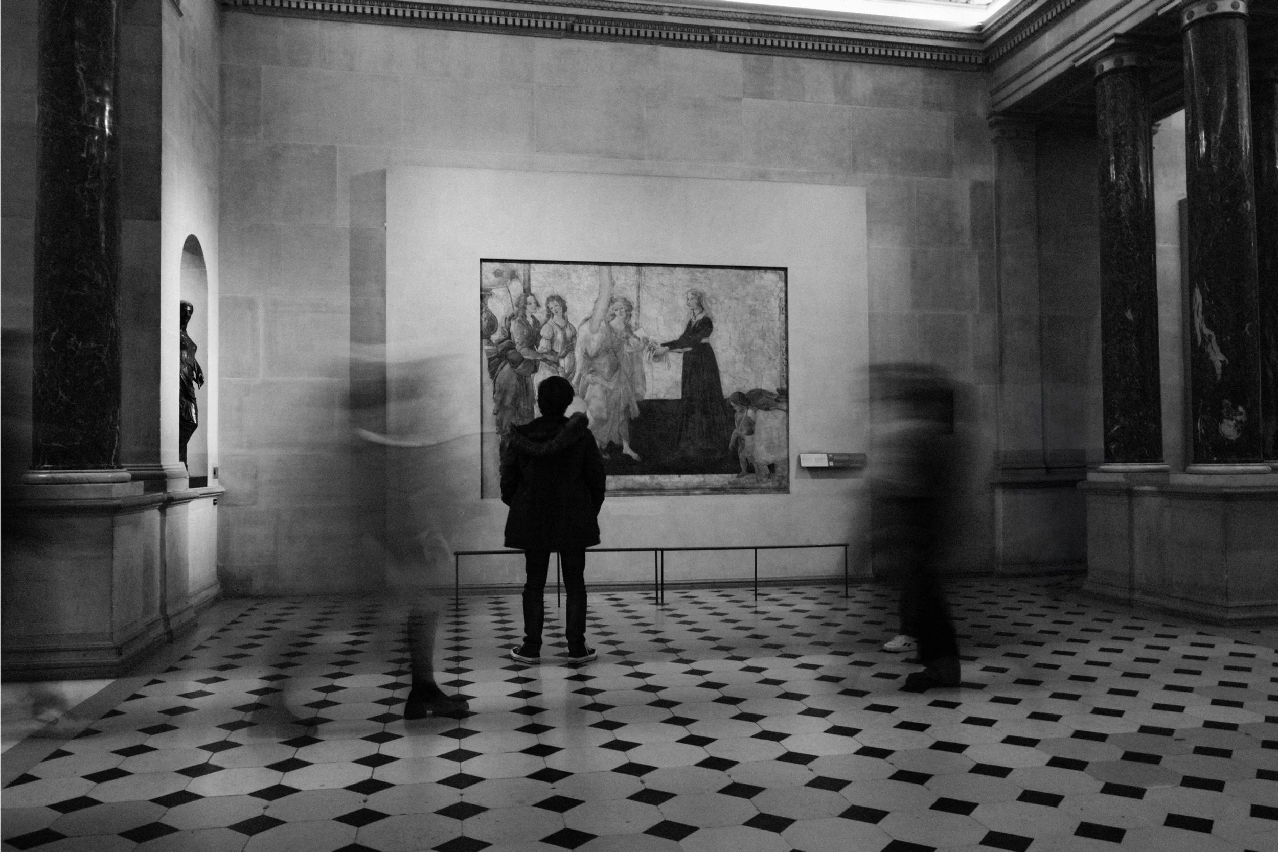 A person standing in an art gallery, looking at a large painting on the wall. The gallery has high ceilings, ornate moldings, and marble columns. The floor is checkered black and white.