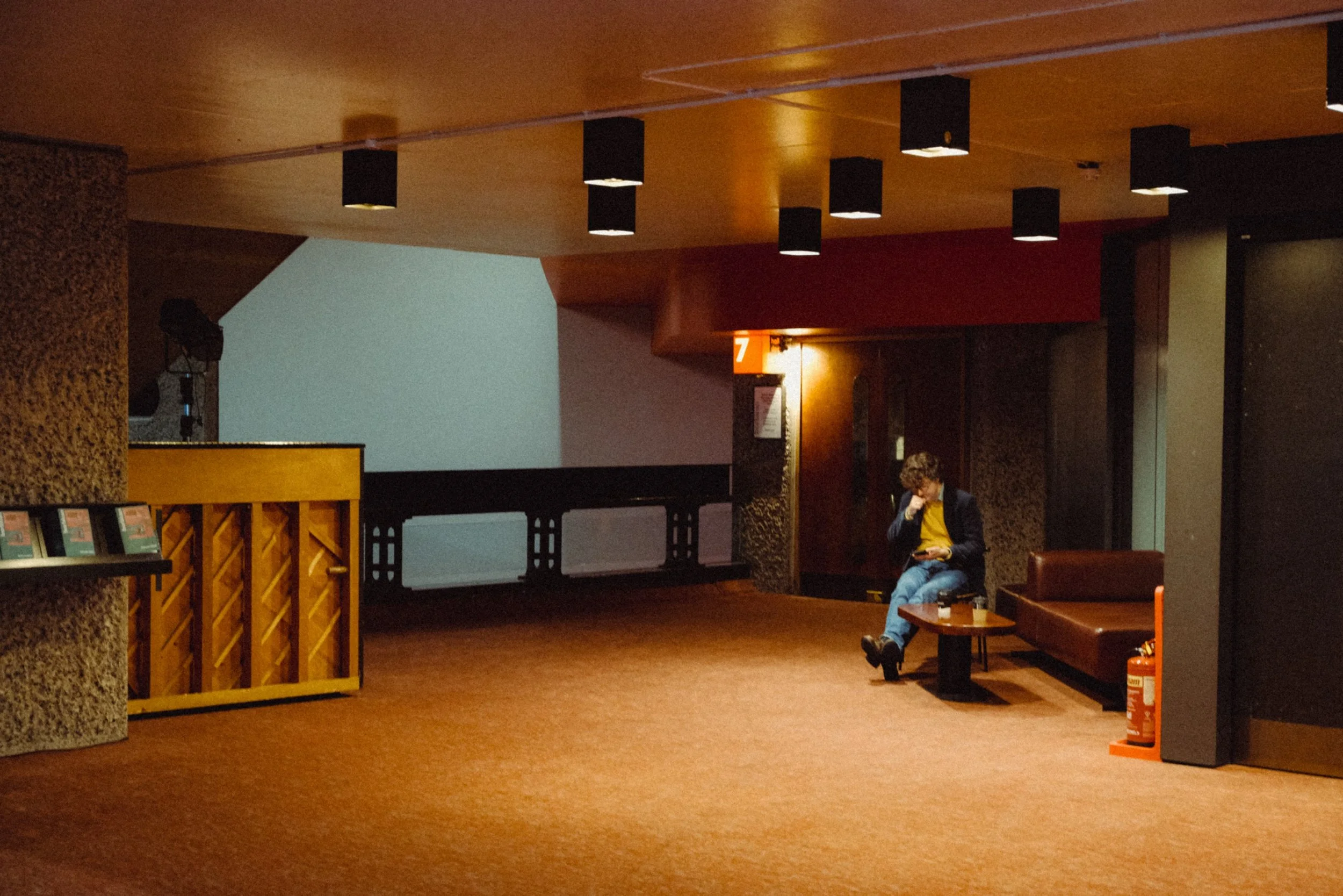 A woman sitting on a leather bench reading a book or phone in a dimly lit indoor waiting area with a wood-paneled ceiling and a red wall with a number 7 sign, and a fire extinguisher on the floor beside her.