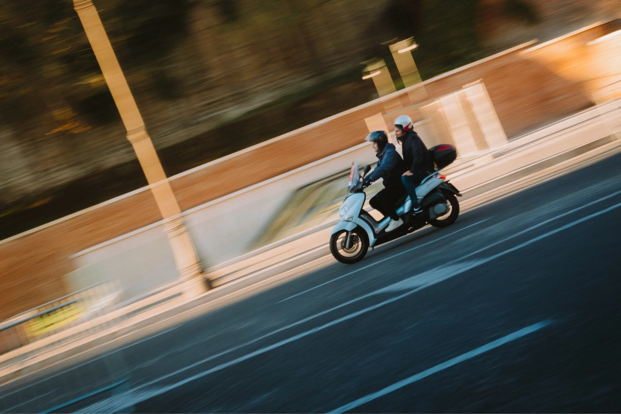 Two people wearing helmets riding a scooter on a road with a fence and trees in the background during sunset