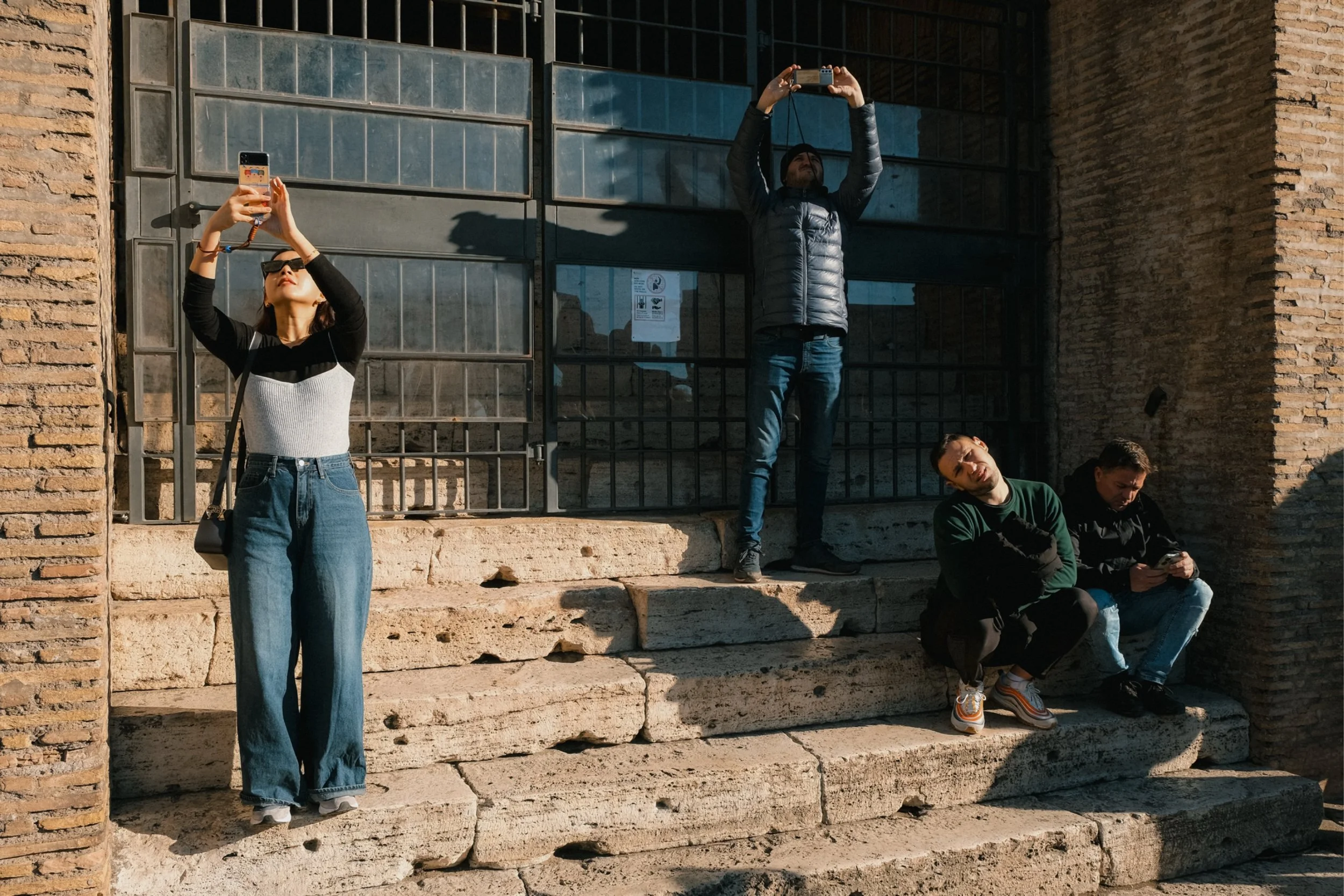 A woman wearing sunglasses and a black long-sleeve top with a white ribbed tank top over it takes a selfie with her phone. Behind her, a man in a black puffer jacket and beanie stands on steps, holding his phone up to take a picture. Two other men si