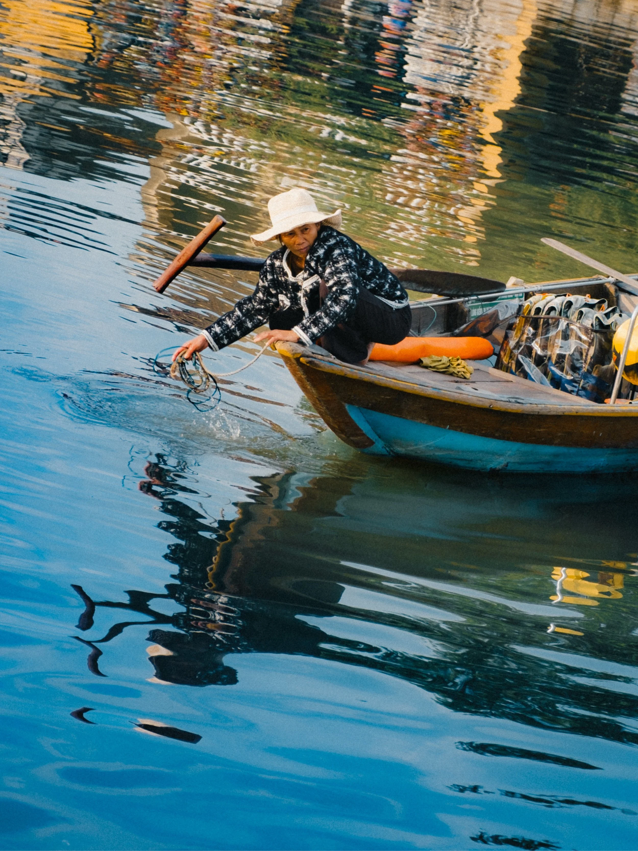 A person in a boat wearing a straw hat and patterned jacket, holding a rope, with reflections of buildings and trees on the water.
