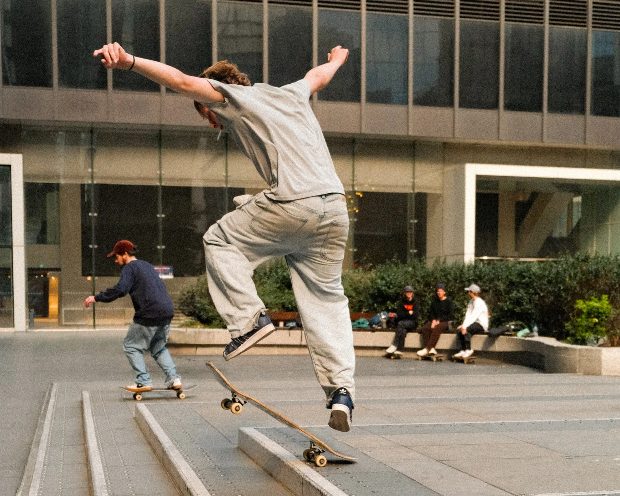 A person skating on a skateboard in an urban skatepark at night, with a group of people sitting on benches in the background.