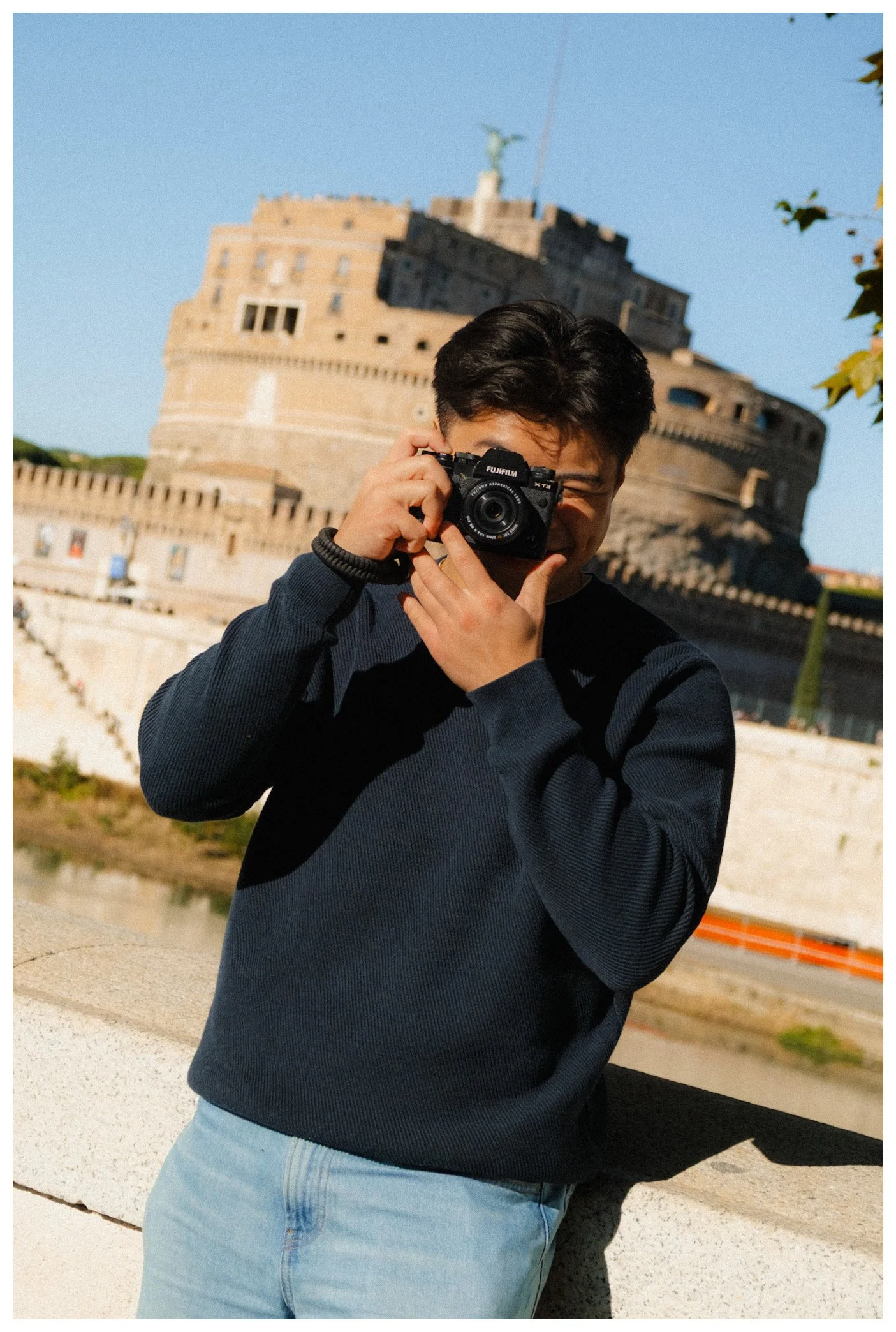 A young man with dark hair holding a camera in front of his face, standing outdoors near a large historic fortress and a river, under a clear blue sky.