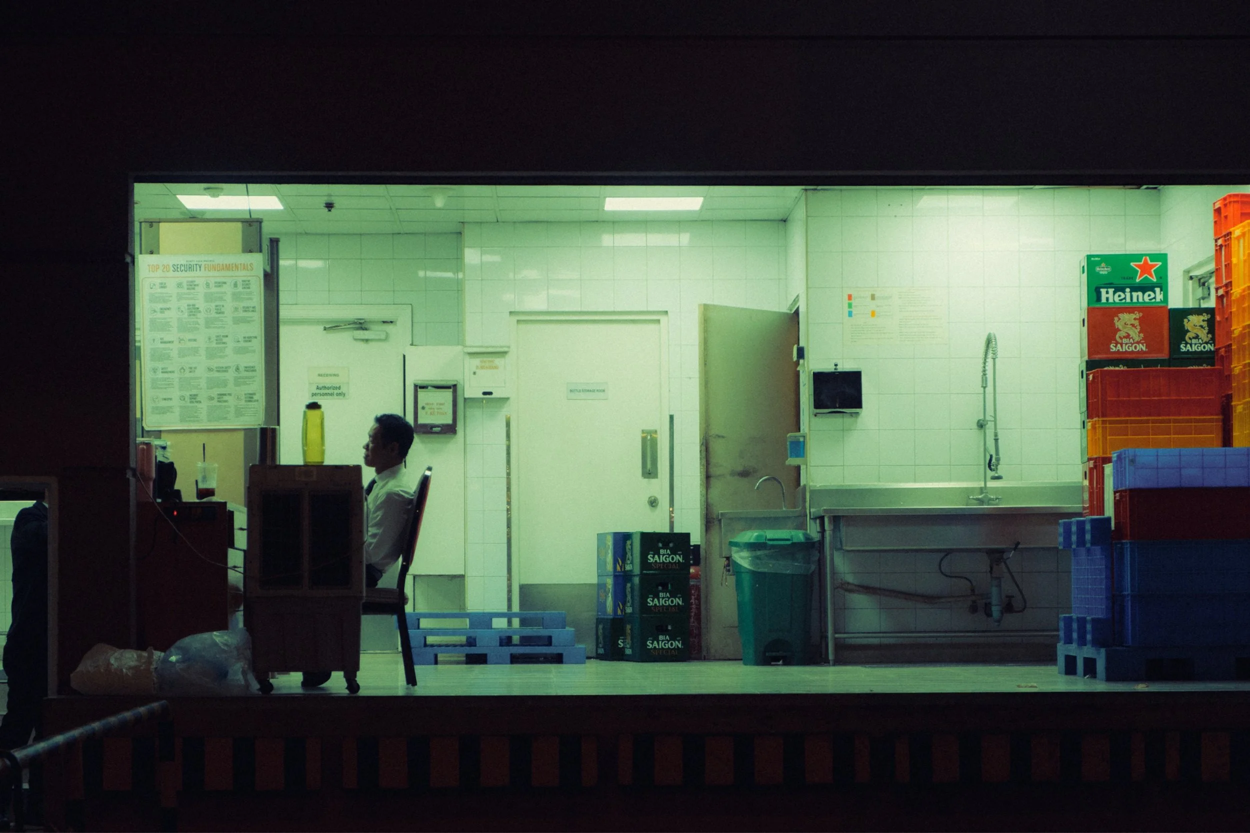 Inside view of a small commercial kitchen with white tiled walls, a sink, stacked crates with Heineken and Saigon beer logos, and a person sitting at a desk near the left side.