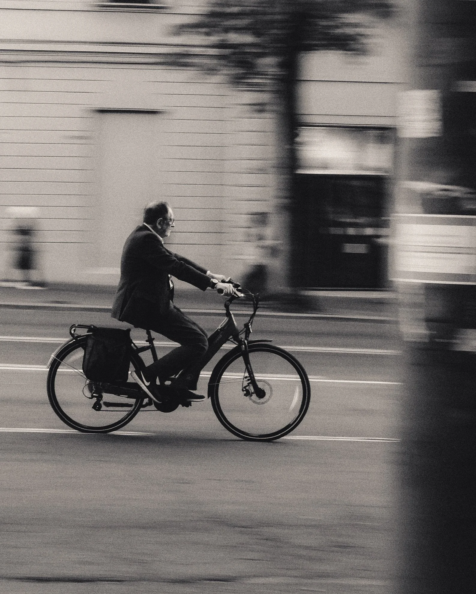 A man wearing glasses and a business suit riding a bicycle on a street. The background is blurred with horizontal lines, suggesting motion.