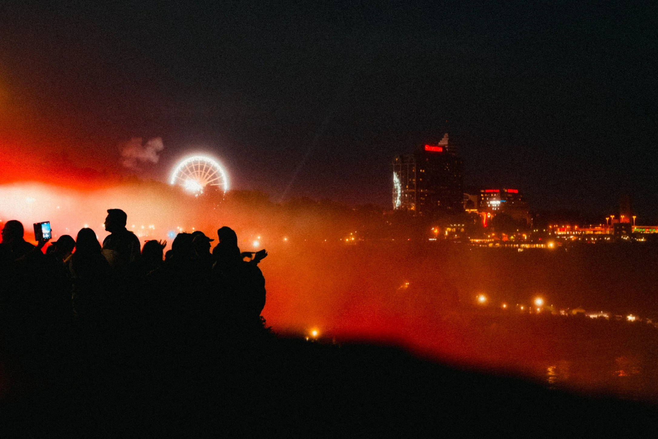 Nighttime cityscape with illuminated ferris wheel, high-rise buildings, and silhouetted people in foreground.
