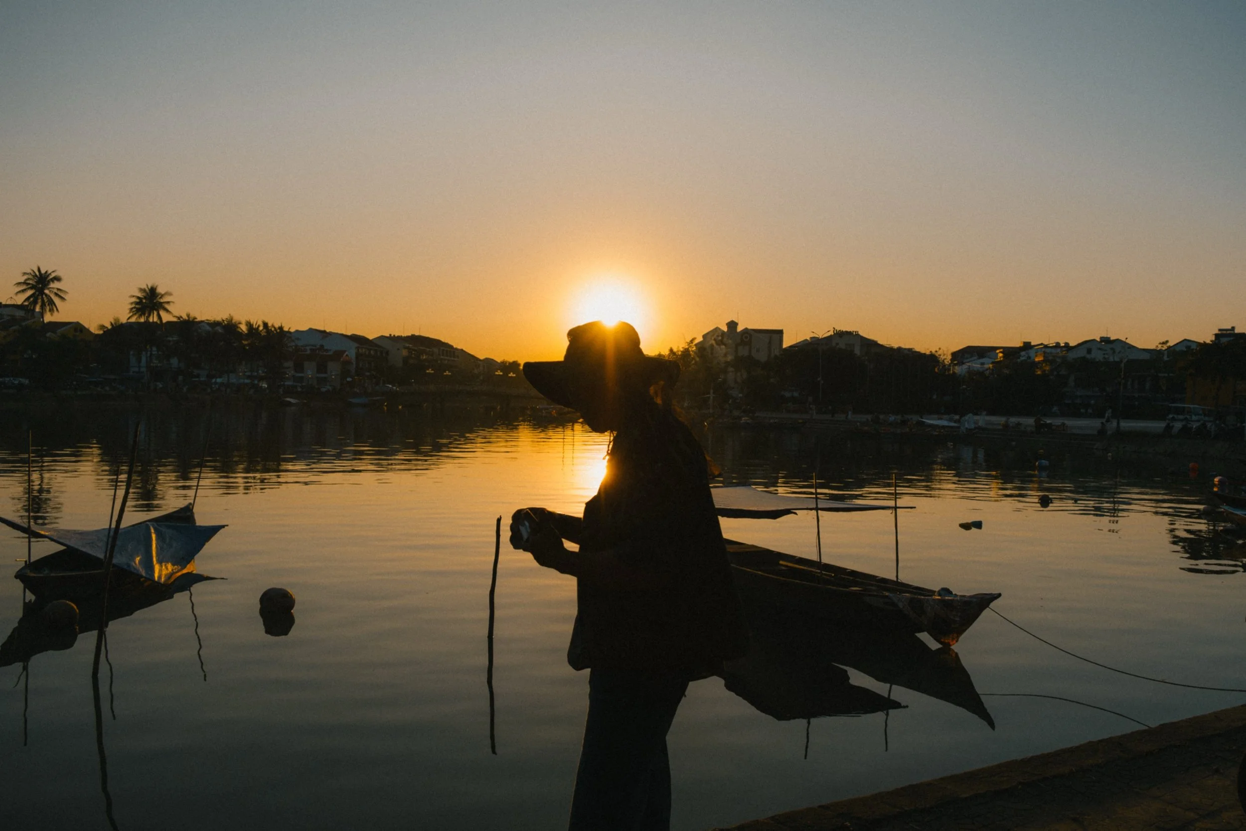 A person at a dock holding a fish, with a sunset behind them and houses and palm trees in the background.