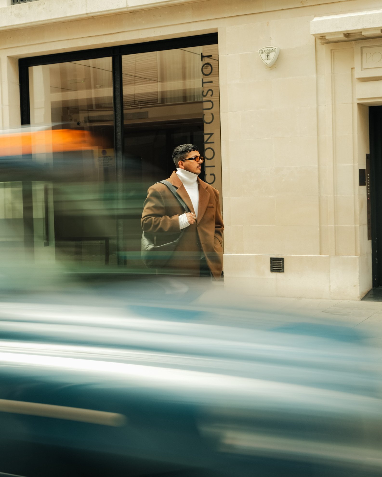 A man with sunglasses and a brown coat stands outside a building with a sign that says 'GION CUSTO'. A blurred vehicle passes by in the foreground.