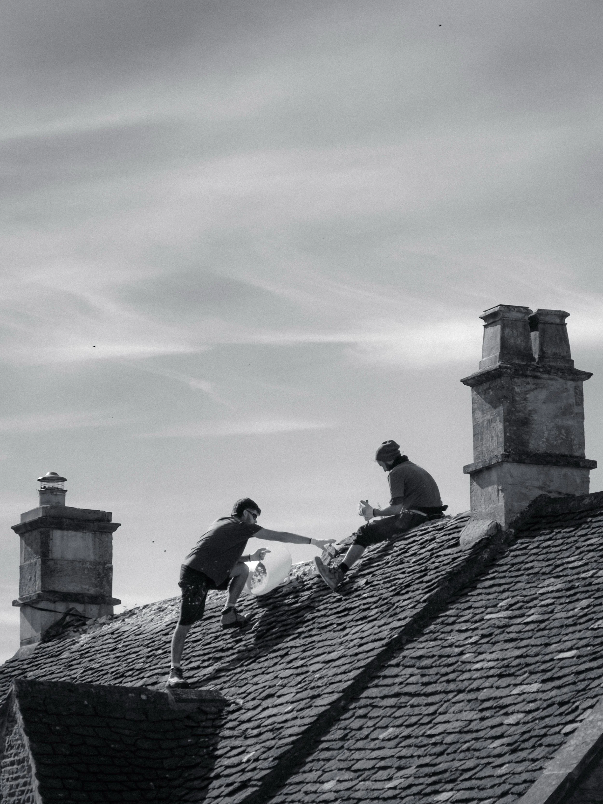 Two people on a rooftop, one handing something to the other, with chimneys and a cloudy sky in the background.