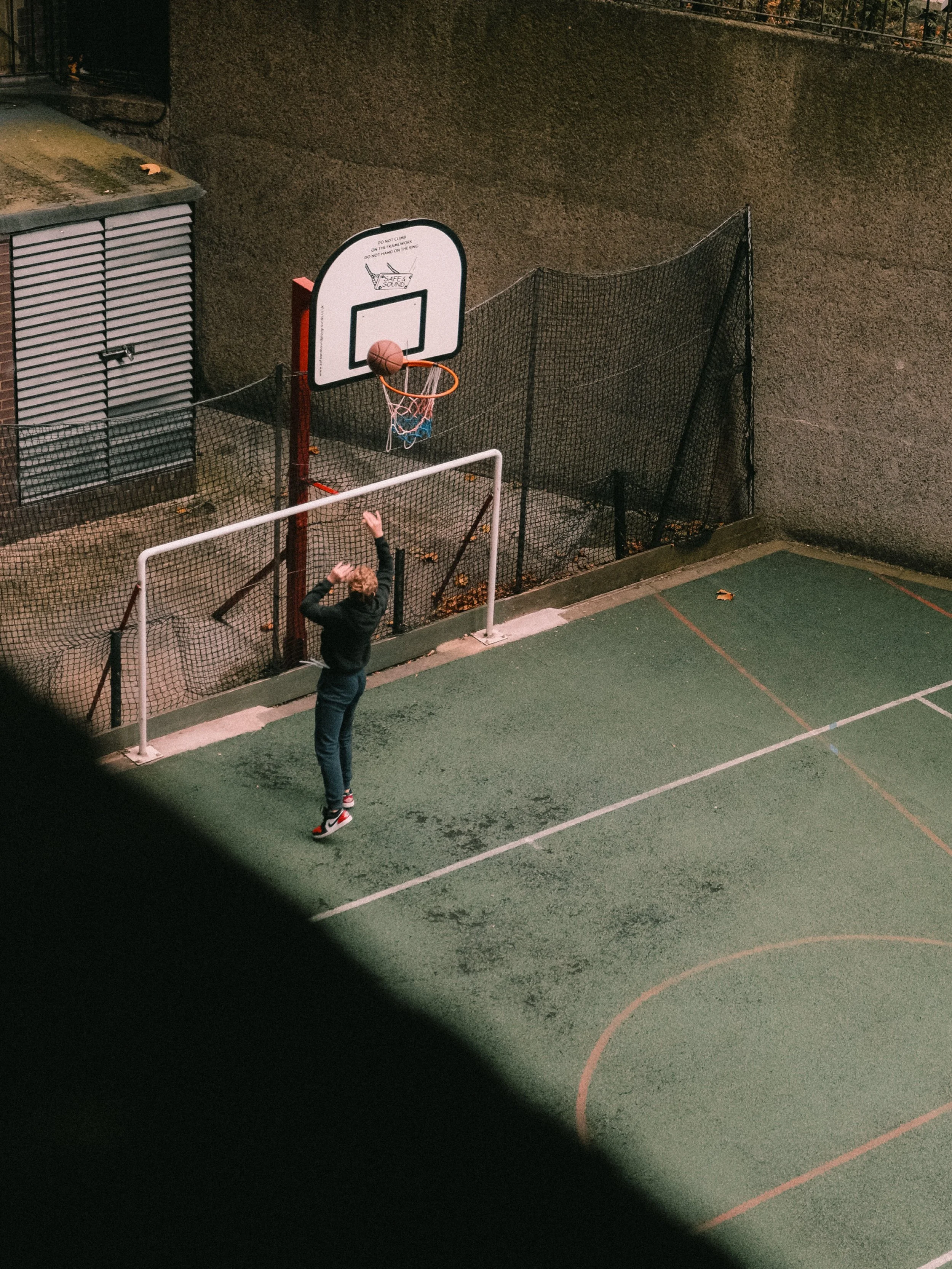 A person standing at a basketball hoop on an outdoor court at night, with a black fence and a wall in the background.