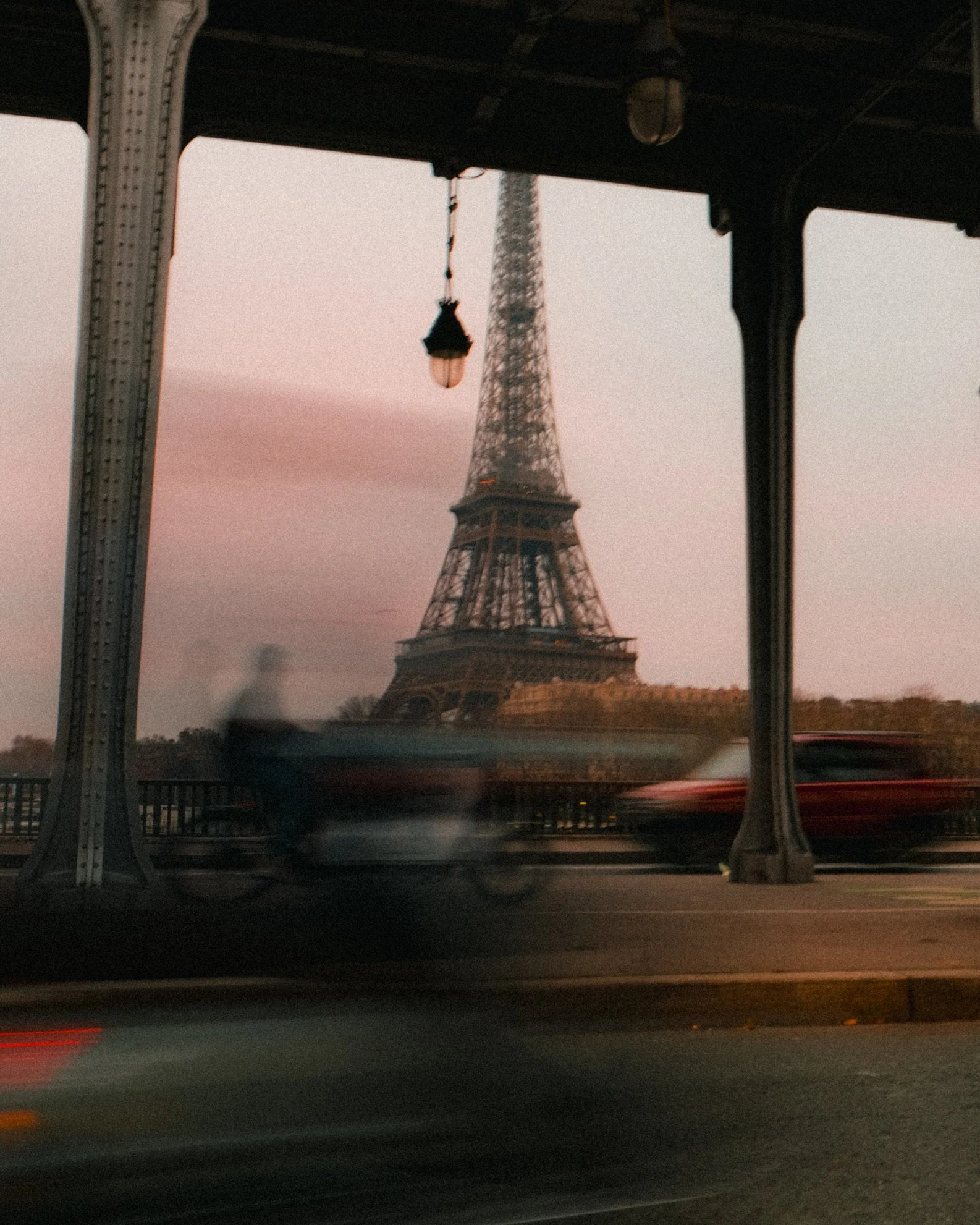 View of the Eiffel Tower through a bridge structure at dusk or dawn, with blurred foreground elements and a pastel sky.