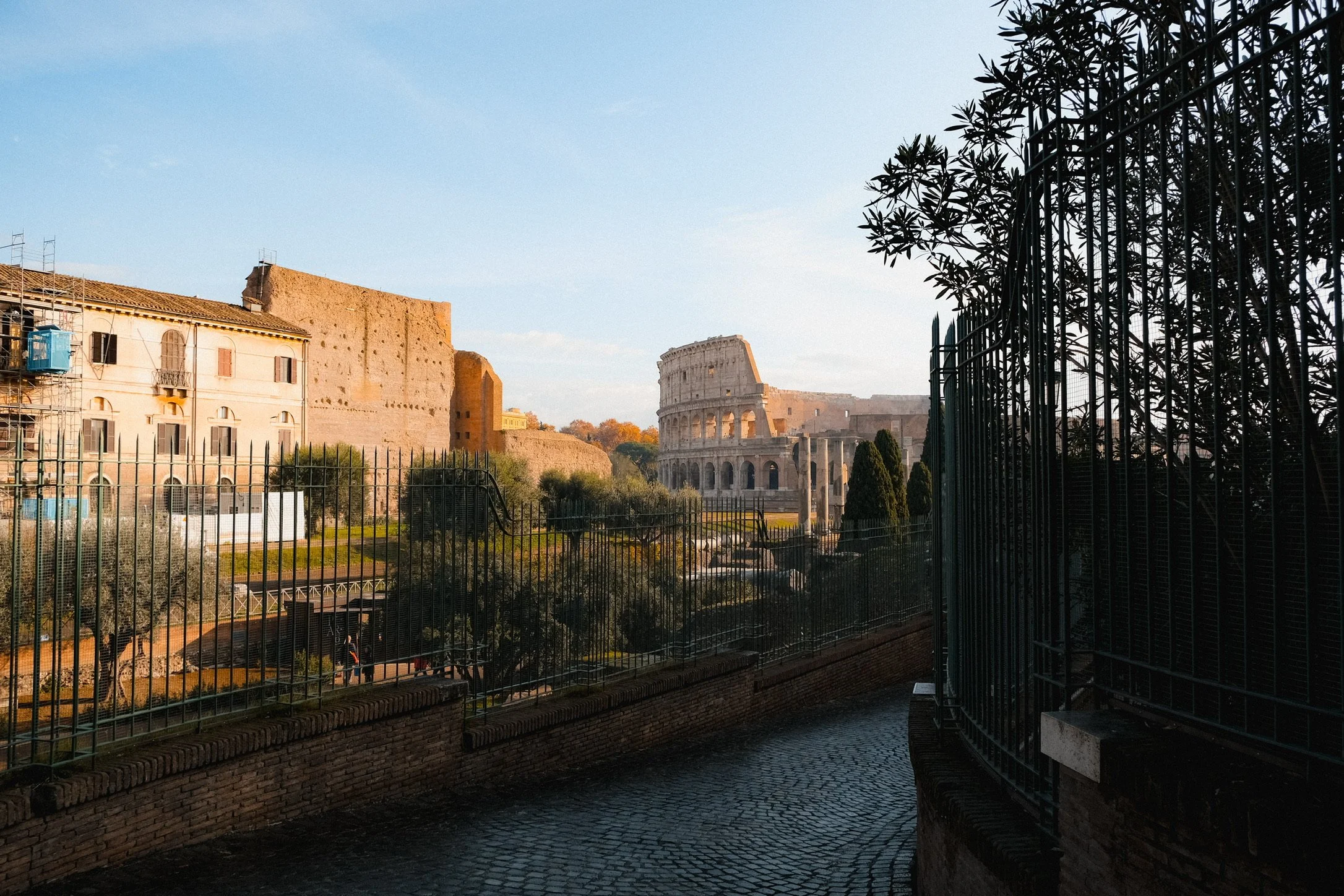 View of the Colosseum and ancient ruins in Rome, Italy, seen through a black metal fence and stone pathway, illuminated by soft sunlight.