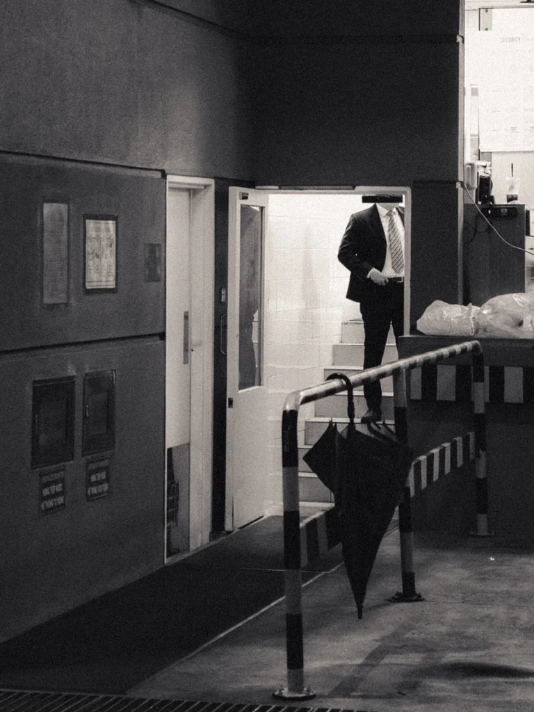 A person in a suit and tie standing at the top of a staircase seen through a doorway in an industrial or commercial setting, with a black umbrella hanging on a striped safety barrier in the foreground.