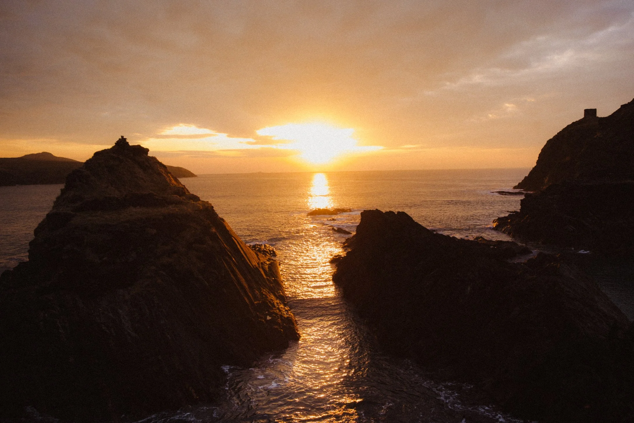 A sunset over the ocean with rocky cliffs on both sides, the sky is partly cloudy with warm orange and yellow hues reflected in the water.