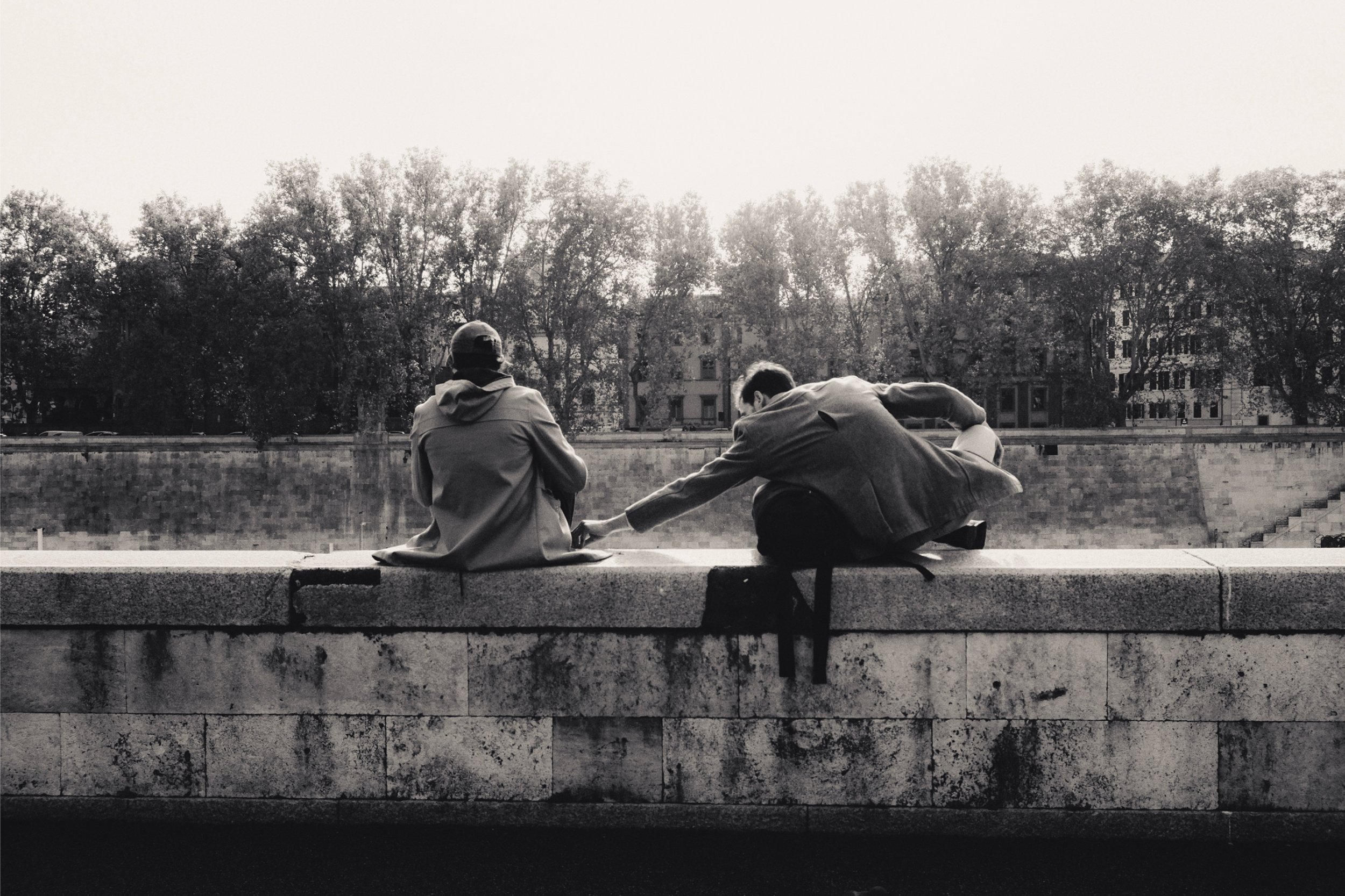 Black and white photo of two men sitting on a stone ledge. One man faces away, wearing a jacket and cap. The other man reclines, extending his arm out to the man facing away. Behind them are trees and residential buildings.