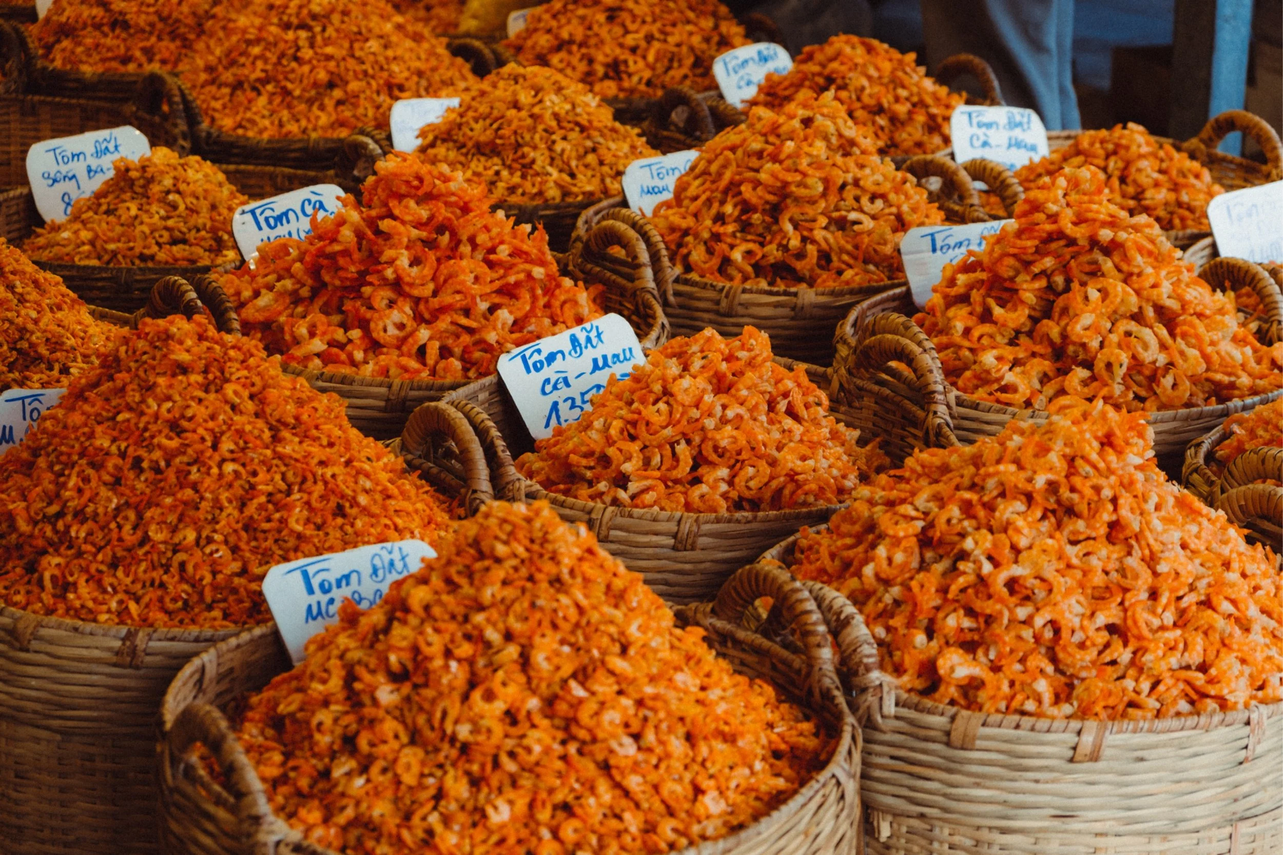 Multiple baskets filled with dried small shrimp on a market stall, with handwritten labels in a foreign language.