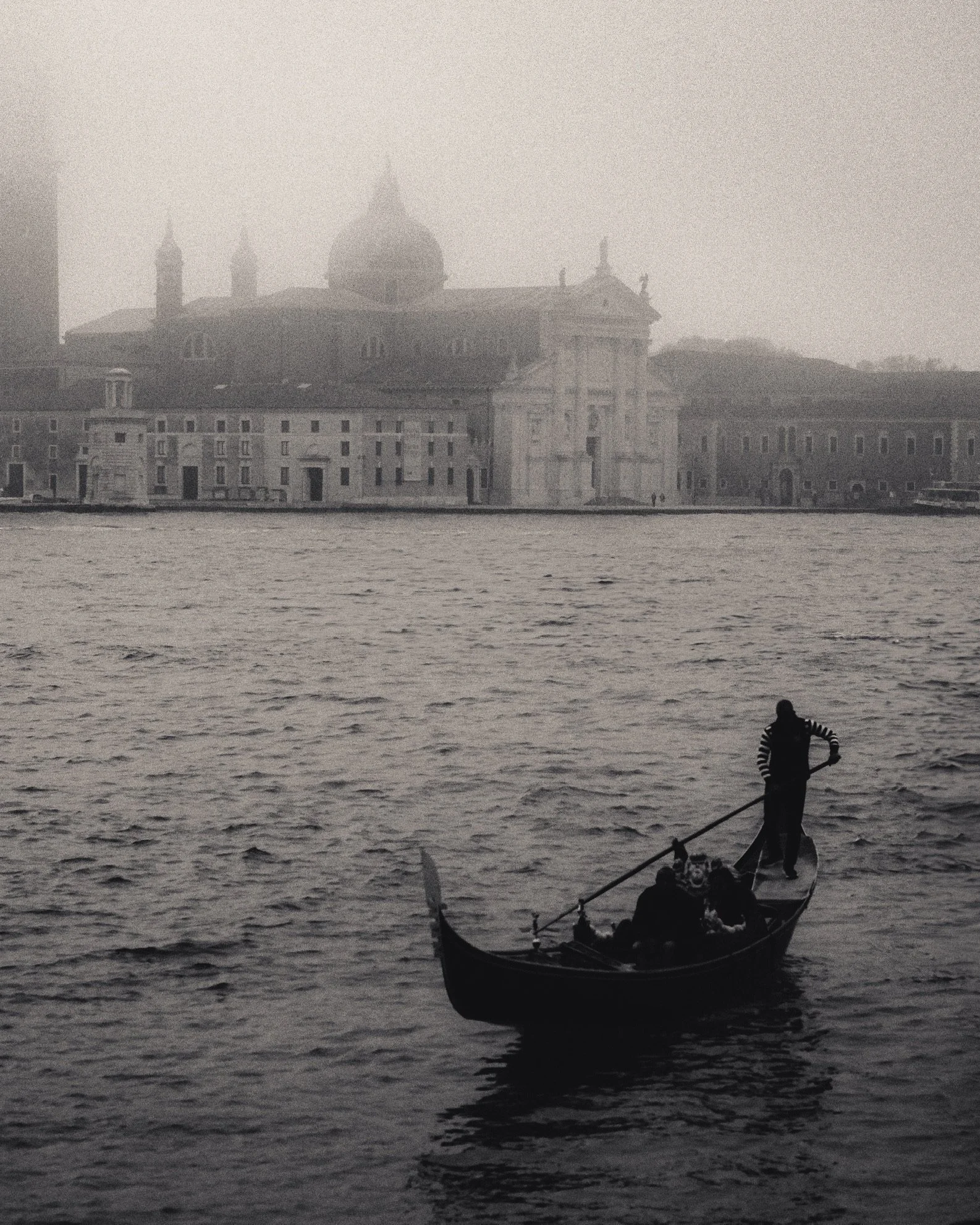 A black and white photo of a person riding a gondola on a body of water, with two dogs sitting in the gondola. In the background, a large historic building with a dome and multiple towers can be seen through mist or fog.