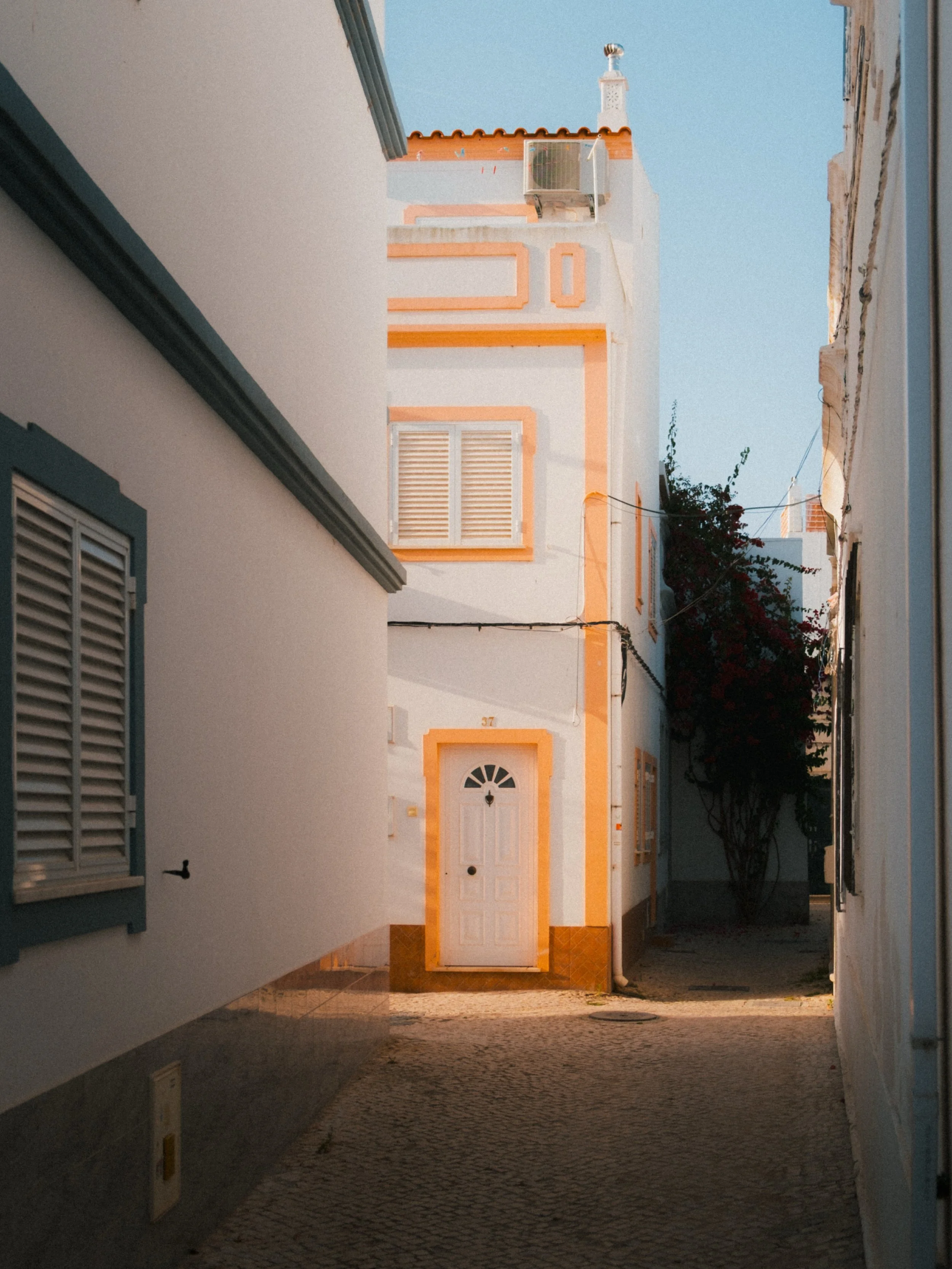 Narrow street or alleyway between white buildings with window shutters, leading to a house with a white door and orange trim, under a clear blue sky.