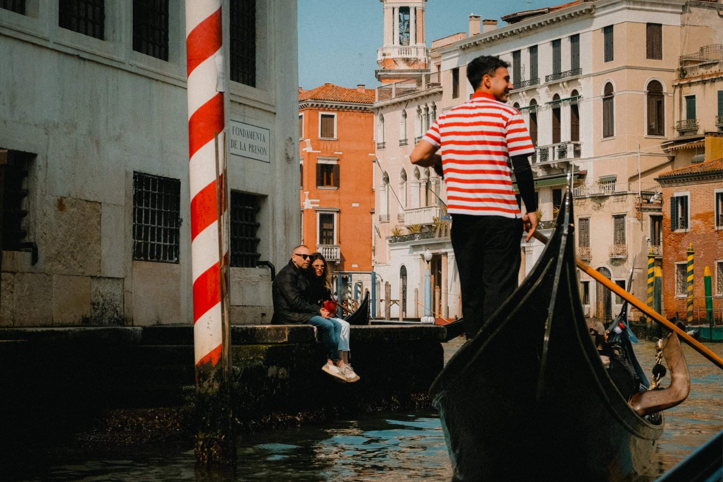 Several people sitting along a canal in Venice, Italy, with colorful historic buildings in the background. A gondola and gondolier are in the foreground.