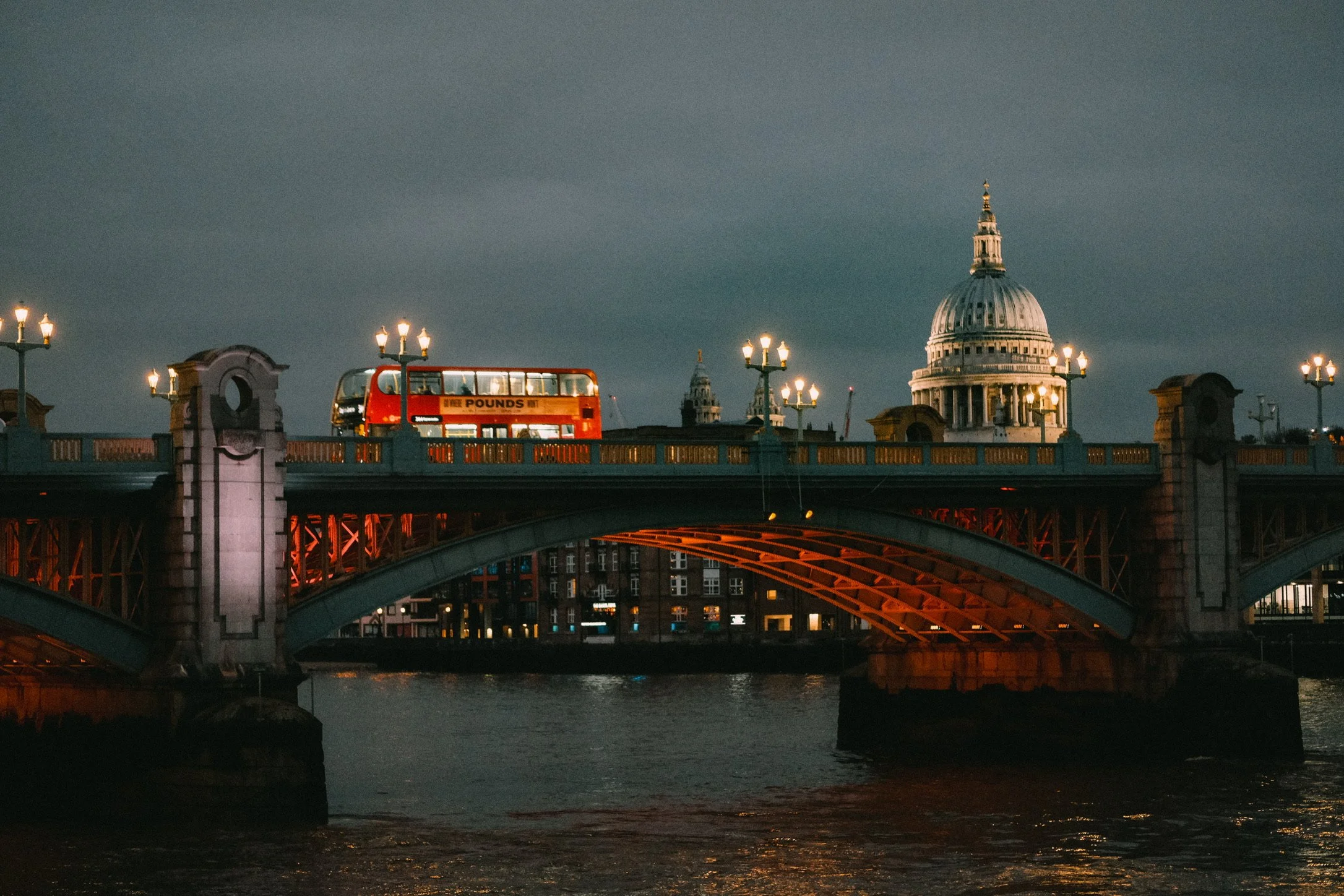 A cityscape at dusk featuring a bridge with traditional street lamps, a red double-decker bus, and the dome of St. Paul's Cathedral in London, England.