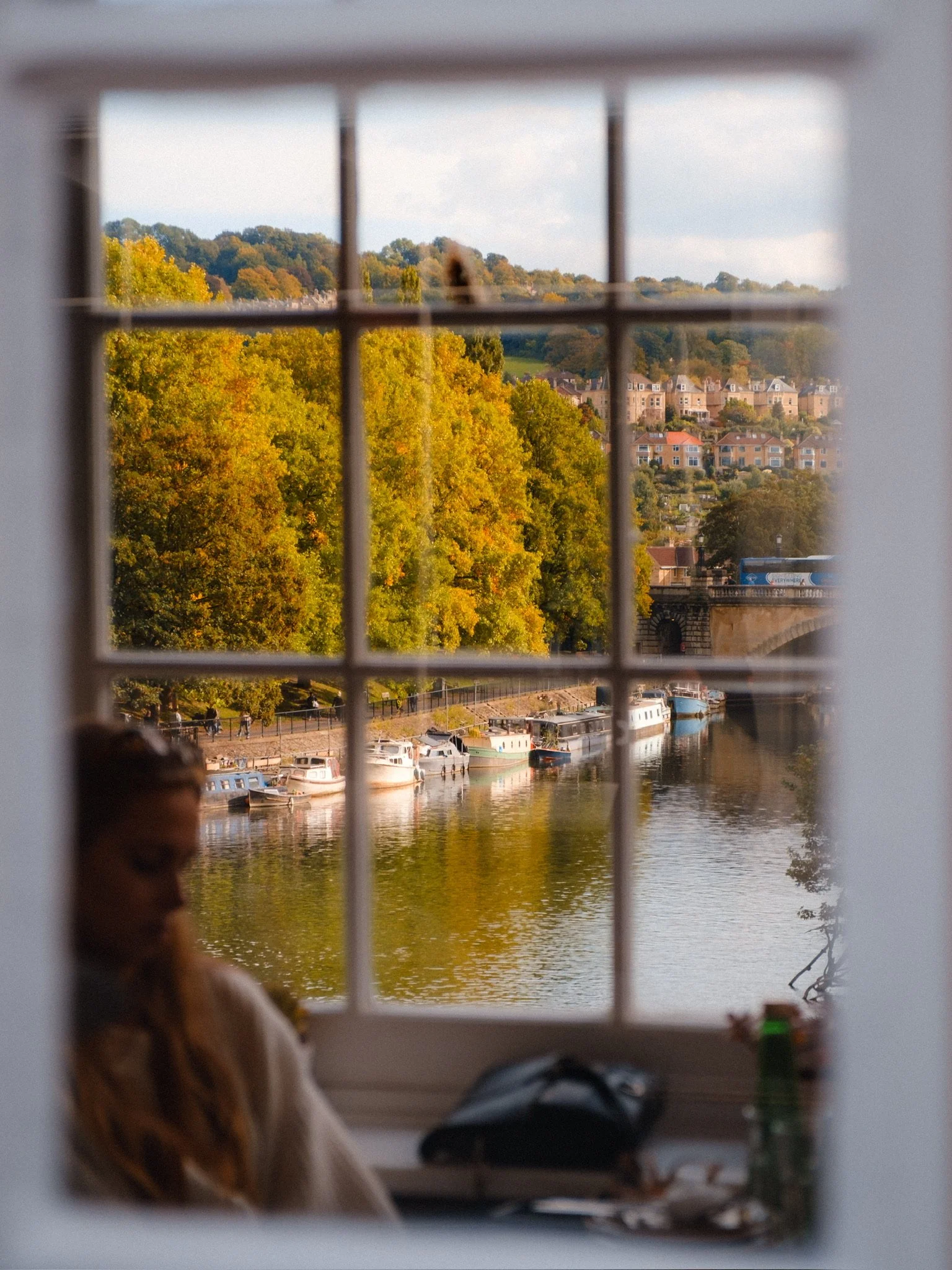 View of a river with boats, trees with fall foliage, and a hillside with houses, seen through a window with grid-like panes.