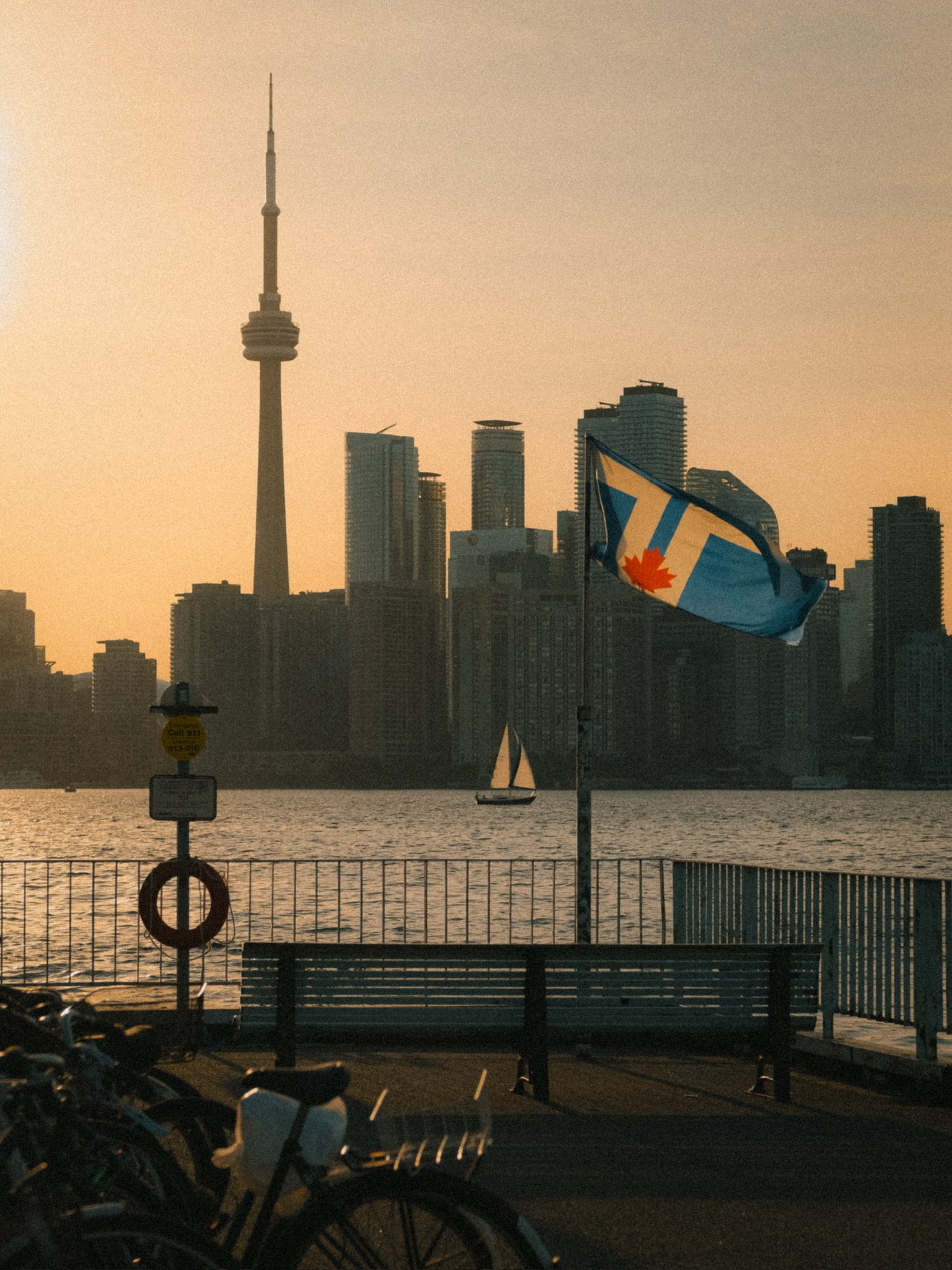City skyline with tall buildings, CN Tower, sailboat on water, Canadian flag, sunset, benches, and bicycles in foreground.