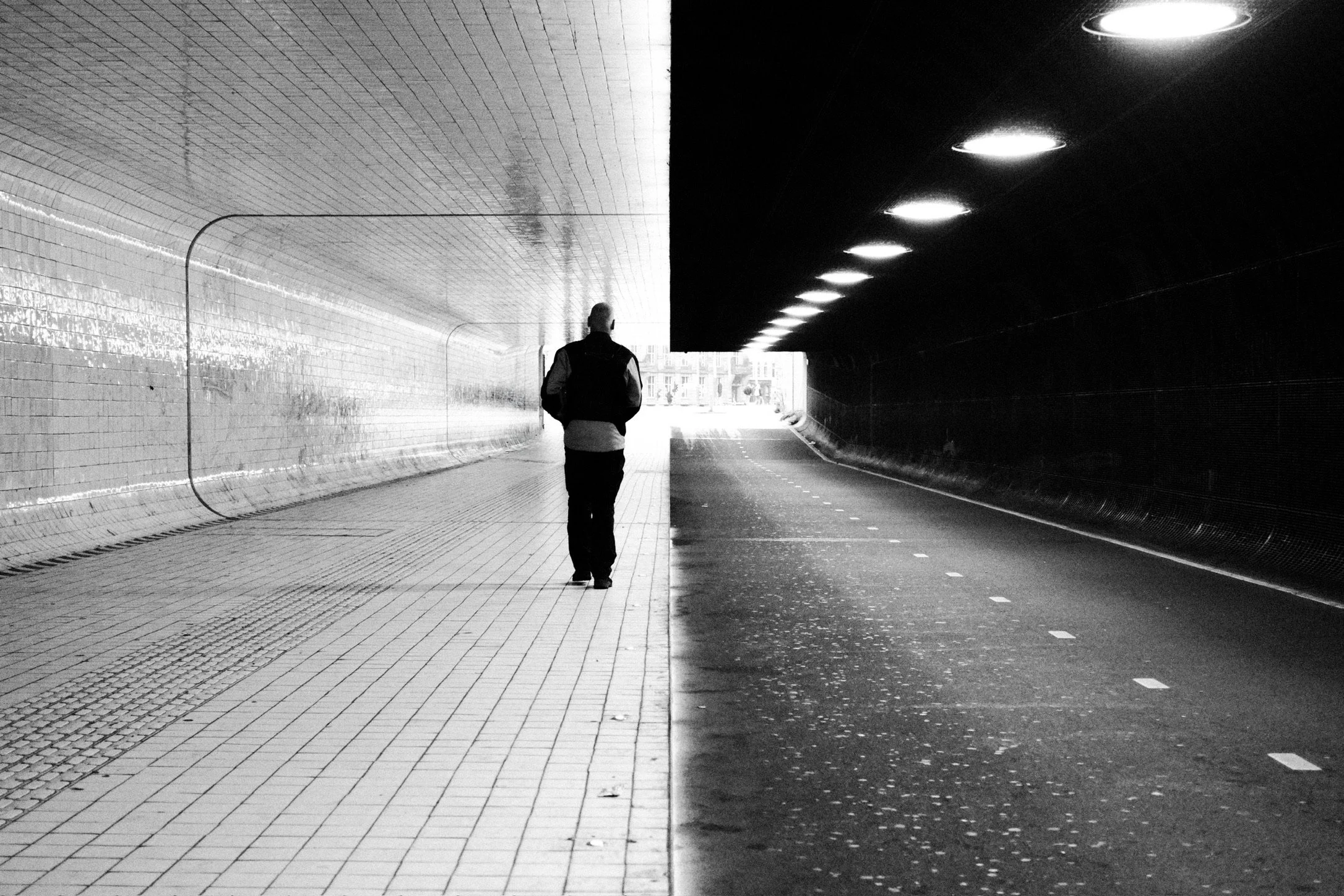 A person walking through a tunnel with contrasting lighting and textures. The left side of the tunnel has tiled walls and floor with a reflective surface, while the right side has dark walls with circular ceiling lights. The image is black and white,
