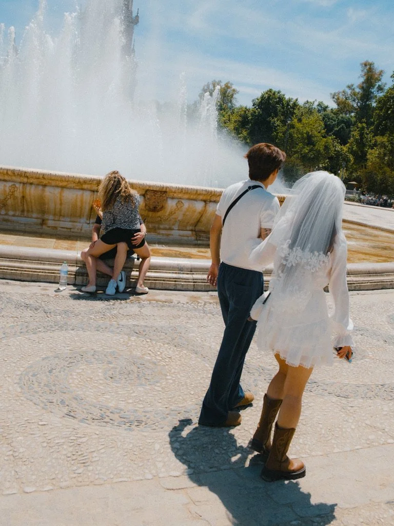 People standing near a fountain in an outdoor park, with one person dressed as a bride and another as a groom, and others sitting on the fountain edge.