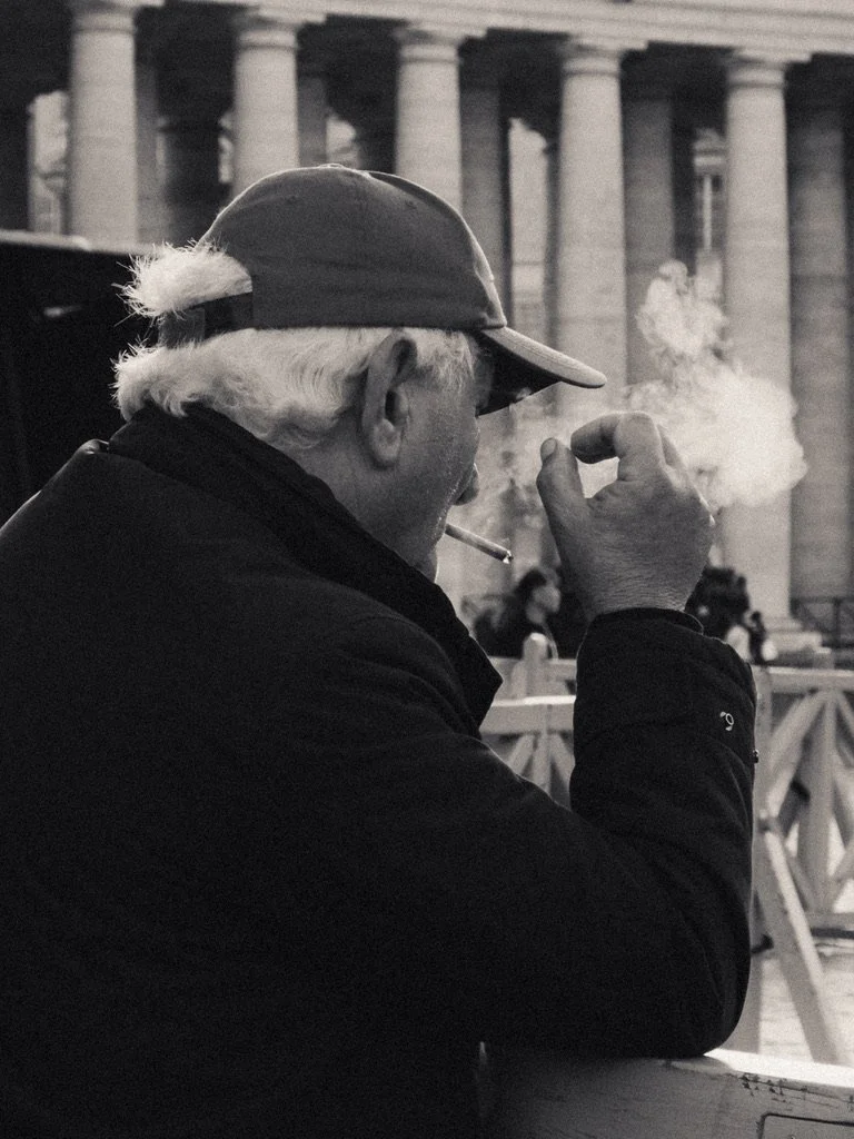 An elderly man with white hair, wearing a baseball cap and glasses, is sitting outdoors in front of a building with large columns. He is smoking a cigarette, and smoke is visible in the image.