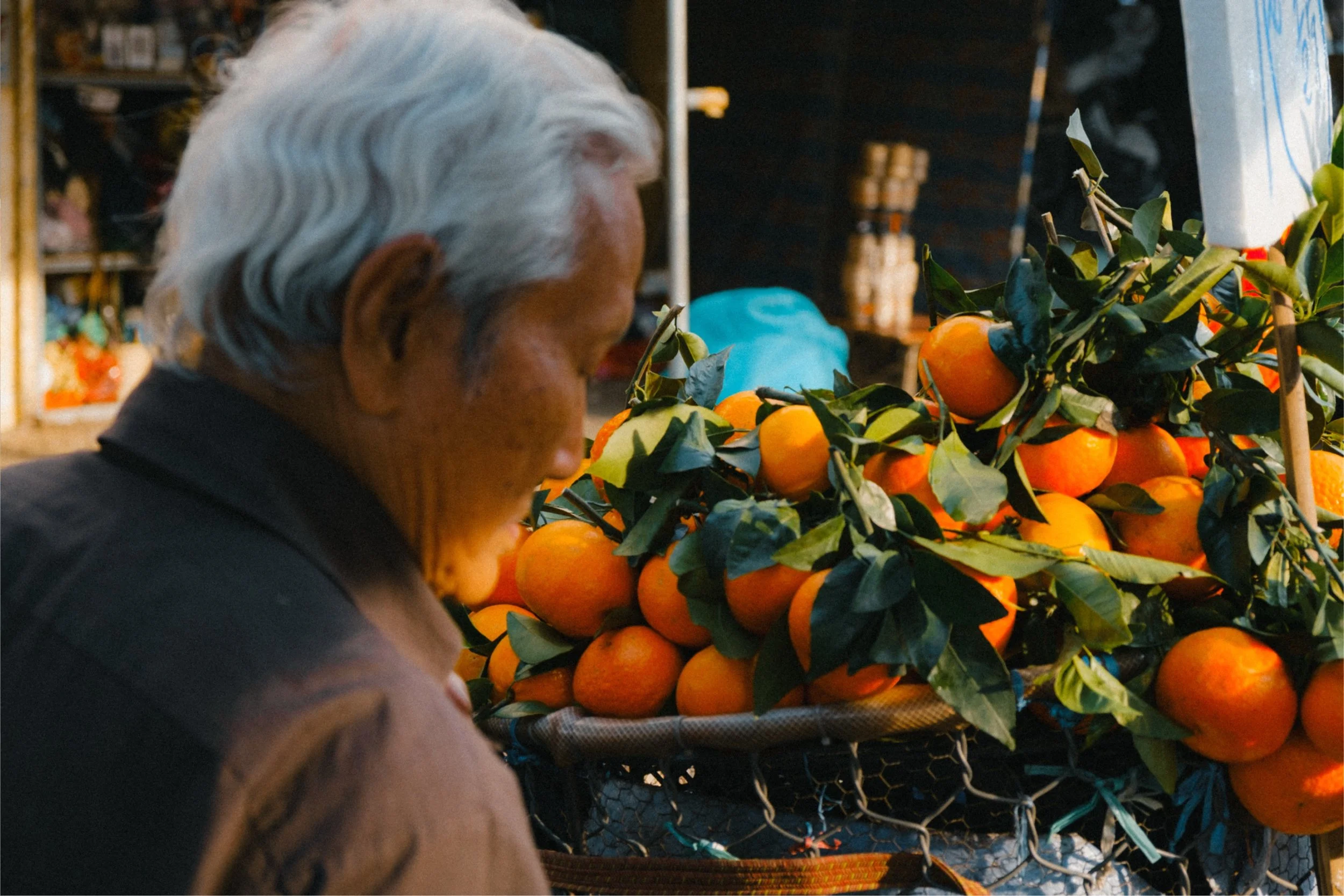 An elderly person with white hair looking at a display of oranges, with a shop or market setting in the background.