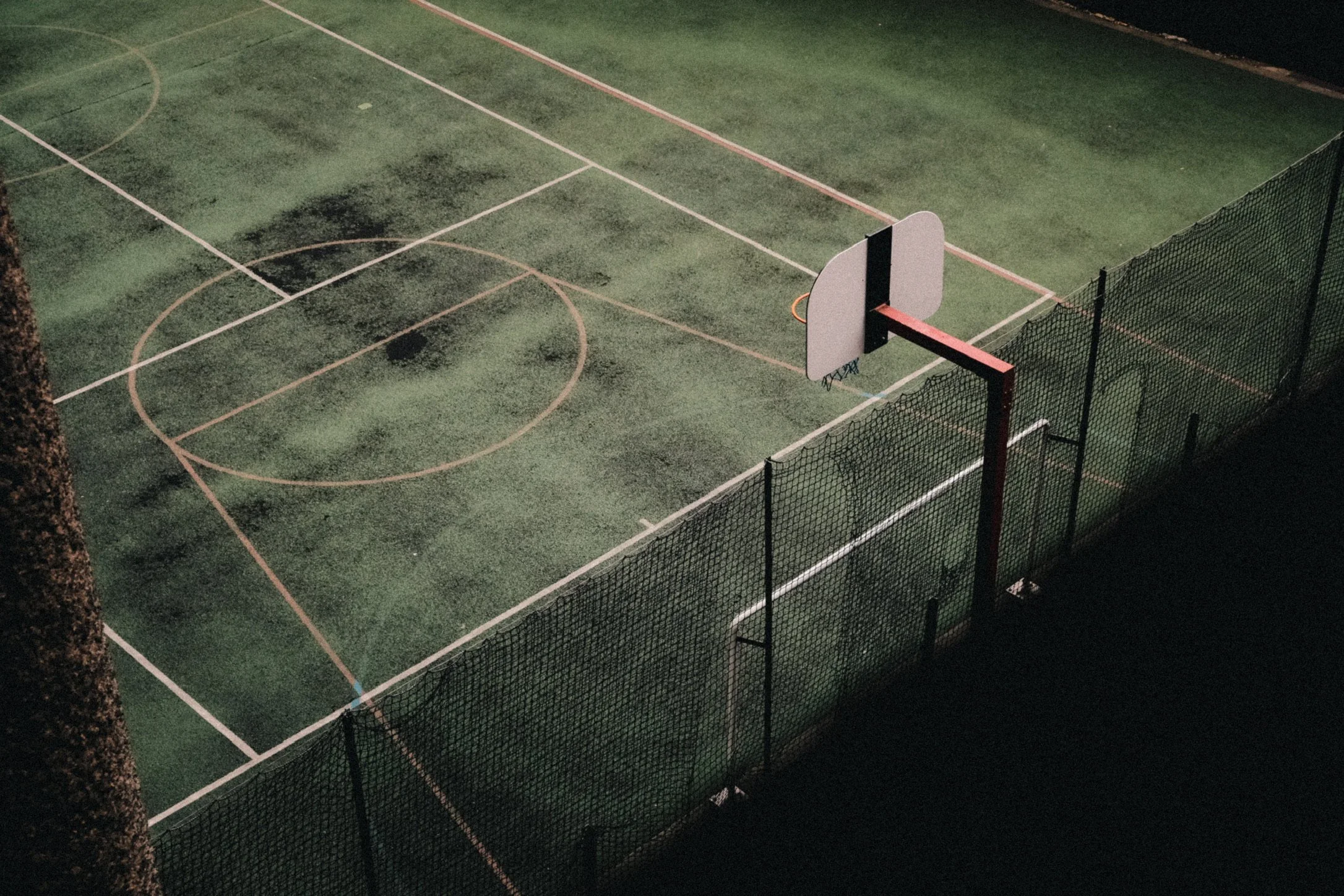 Empty outdoor basketball court at night with a basketball hoop and a chain-link fence.