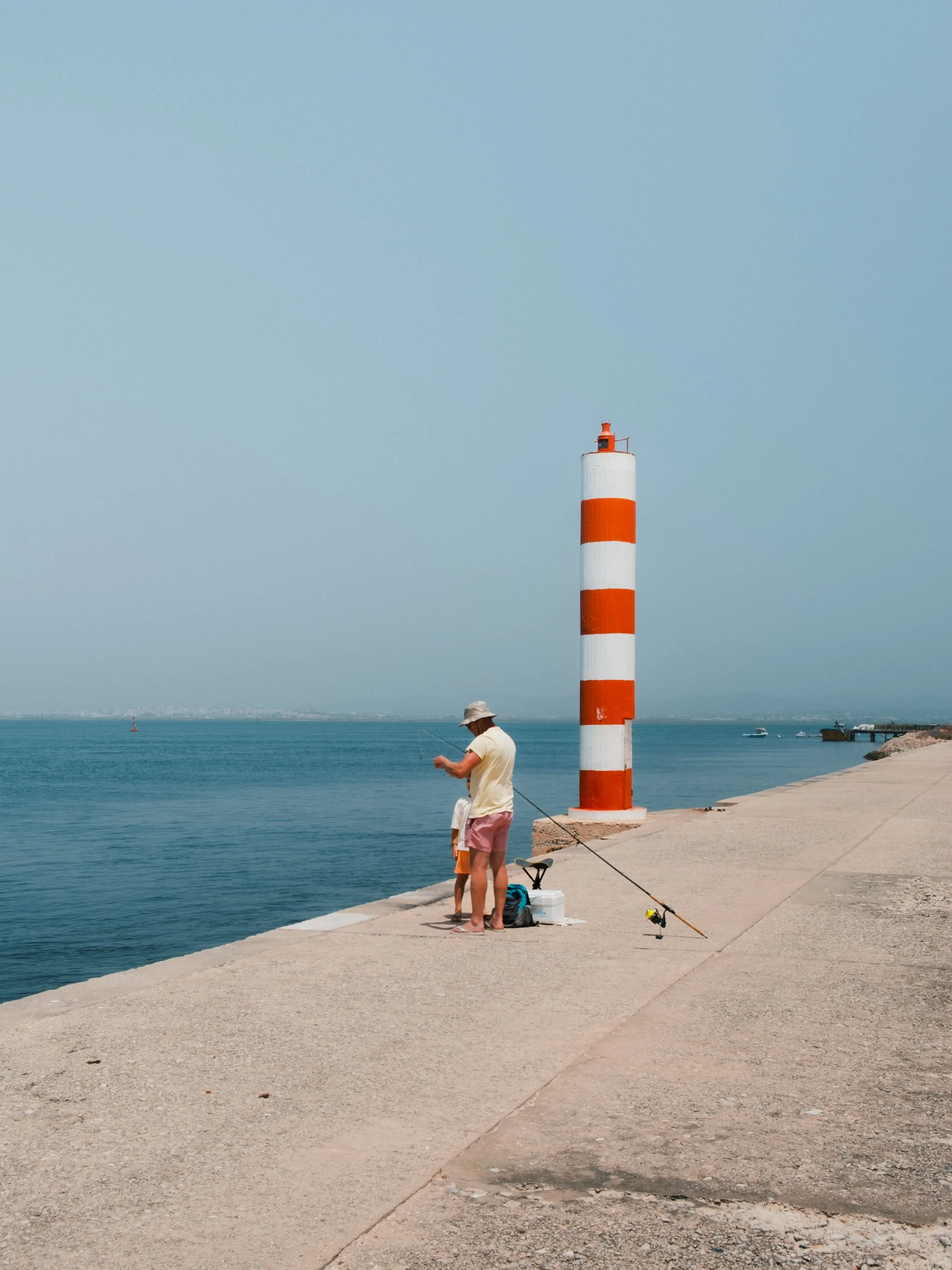 A man in a beige hat and yellow shirt fishing by the waterfront, with a striped orange and white lighthouse nearby and boats visible on the horizon.