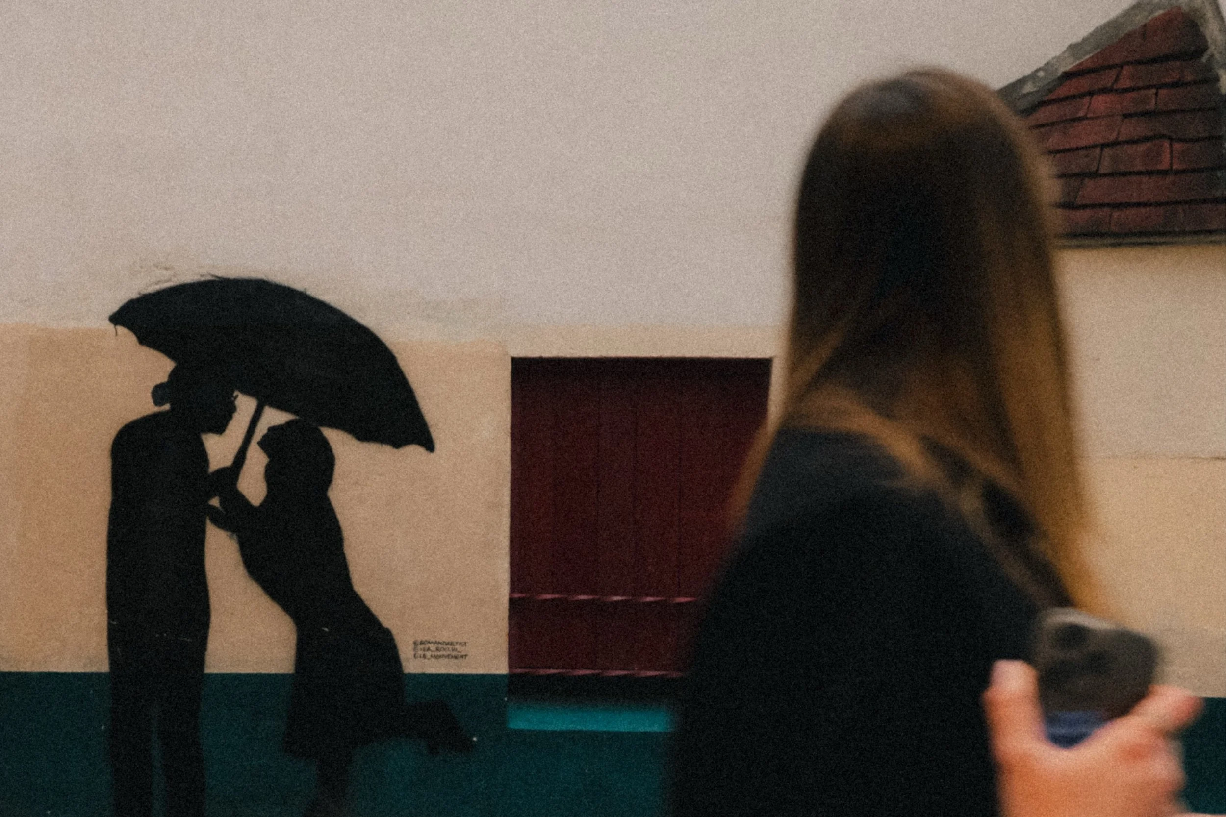 Image of a room with a silhouette wall art piece of two people holding an umbrella. Person in foreground with long hair appears to be walking or running, slightly blurred, with part of their head and arm visible on the right side of the image.