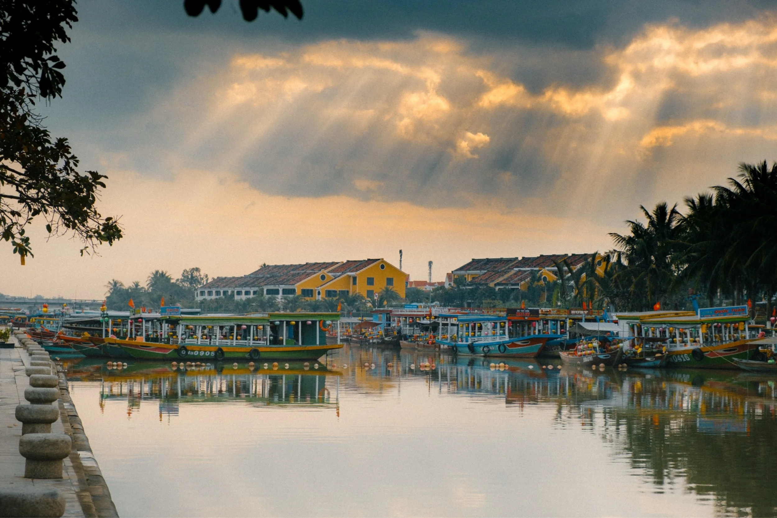 A harbor with colorful boats docked along the water, yellow buildings with tiled roofs in the background, and lush palm trees and other trees along the water's edge. The sky is partly cloudy with sunlight filtering through the clouds.