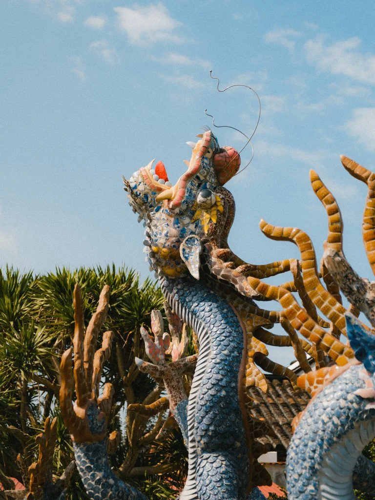 Colorful dragon sculpture with intricate details, surrounded by other artistic figures, set outdoors against a blue sky with some clouds.