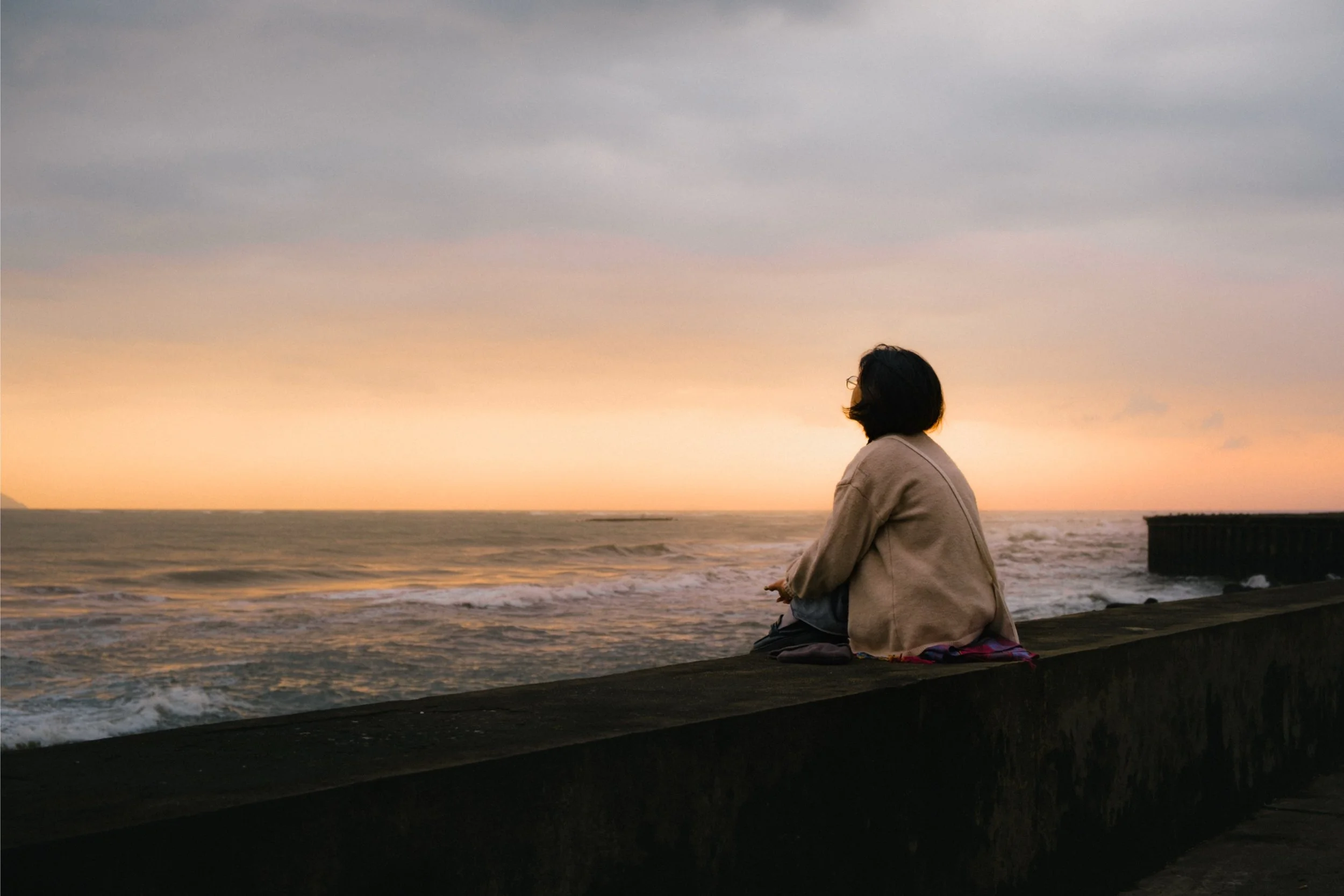 A person with dark hair and glasses, sitting on a beach at sunset, facing the ocean with waves and a sunset sky.