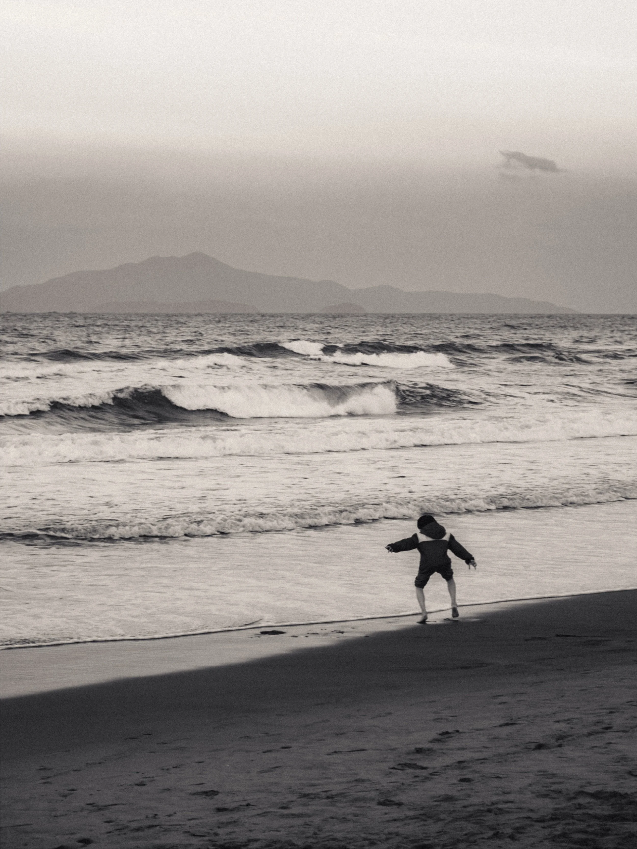 A child standing by the shore at the beach, facing the ocean with waves, and mountains in the background, in black and white.