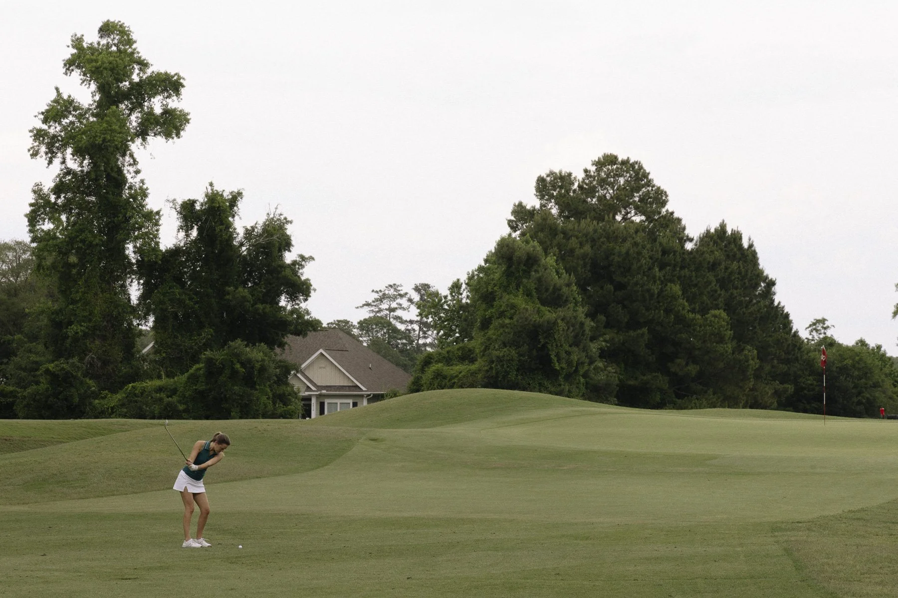 A girl in a teal top and white skirt playing golf on a green golf course, preparing to hit the golf ball with a club, with trees and a house in the background.