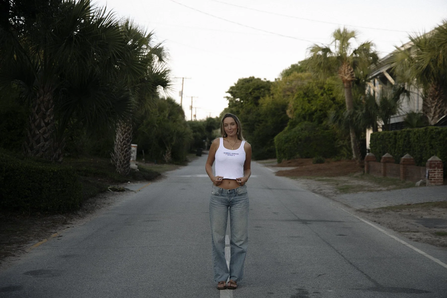 A young woman stands in the middle of a quiet residential street with palm trees and houses on either side. She is wearing a white tank top and baggy jeans, with a smile and her hands in her pockets.