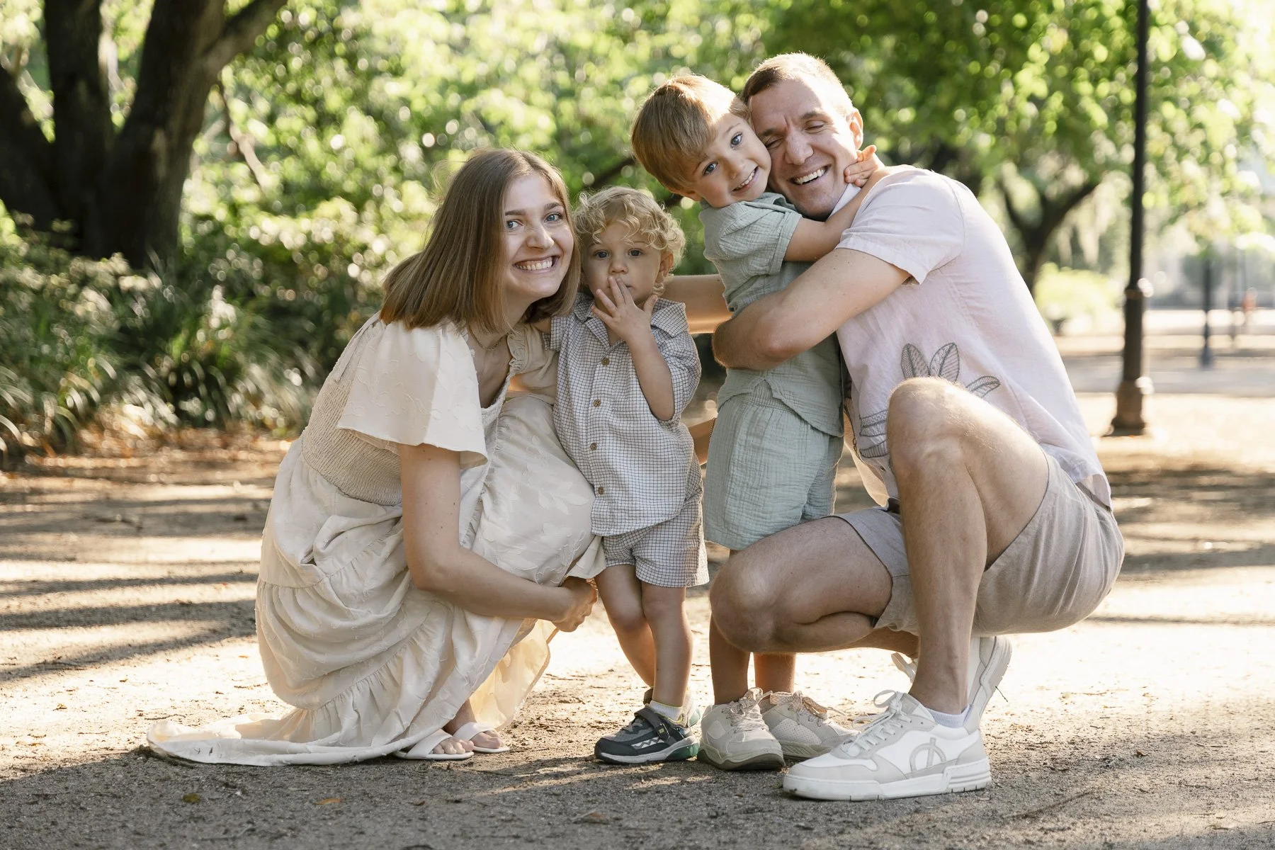 A happy family of four outdoors in a park, smiling and hugging. The mother, father, and two boys are posing together, with lush green trees in the background.
