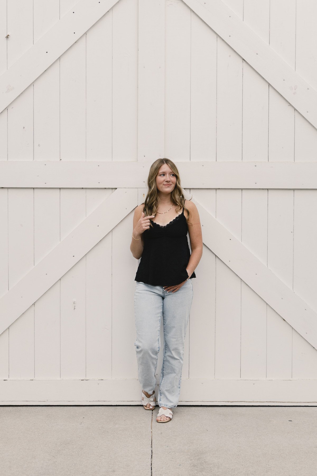 A young woman with wavy brown hair stands in front of a large white barn door with diagonal and horizontal wooden planks. She wears a black lace-trimmed sleeveless top, light blue jeans, and beige sandals. She is looking slightly to her left with a thoughtful expression.