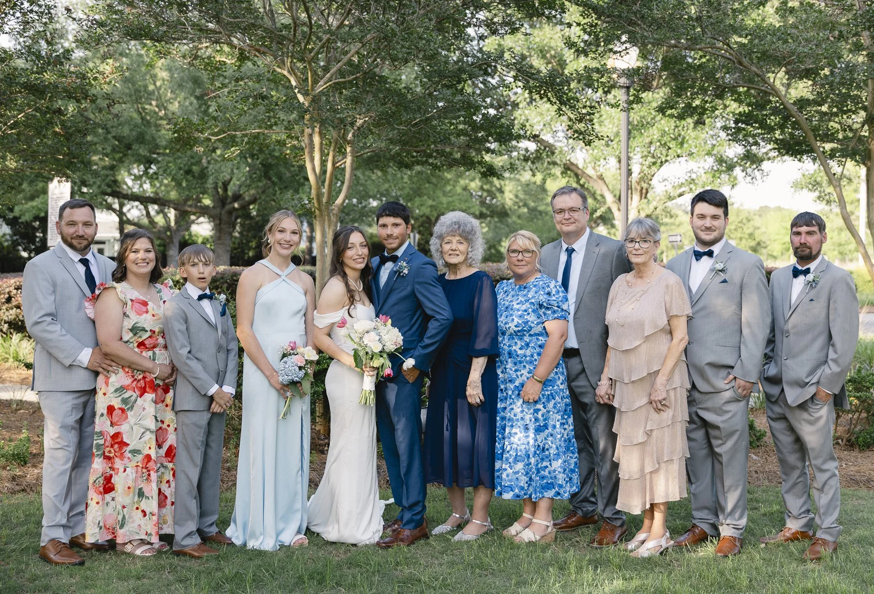 A group of people dressed in formal attire, standing outdoors under trees for a wedding photo. The bride and groom are in the center, smiling and holding flowers, surrounded by family and friends.