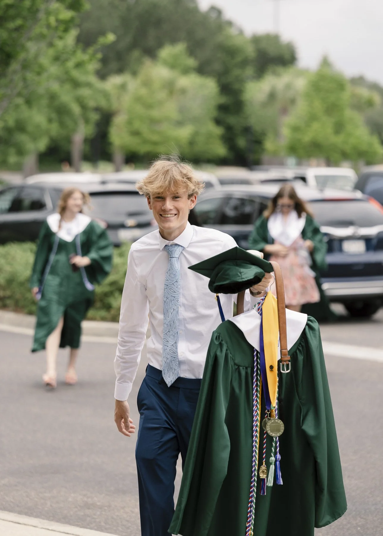 A young man dressed in a white shirt and blue tie, smiling and holding a graduation cap and gown, walking outdoors with cars and trees in the background. Two women in green graduation gowns are walking behind him.