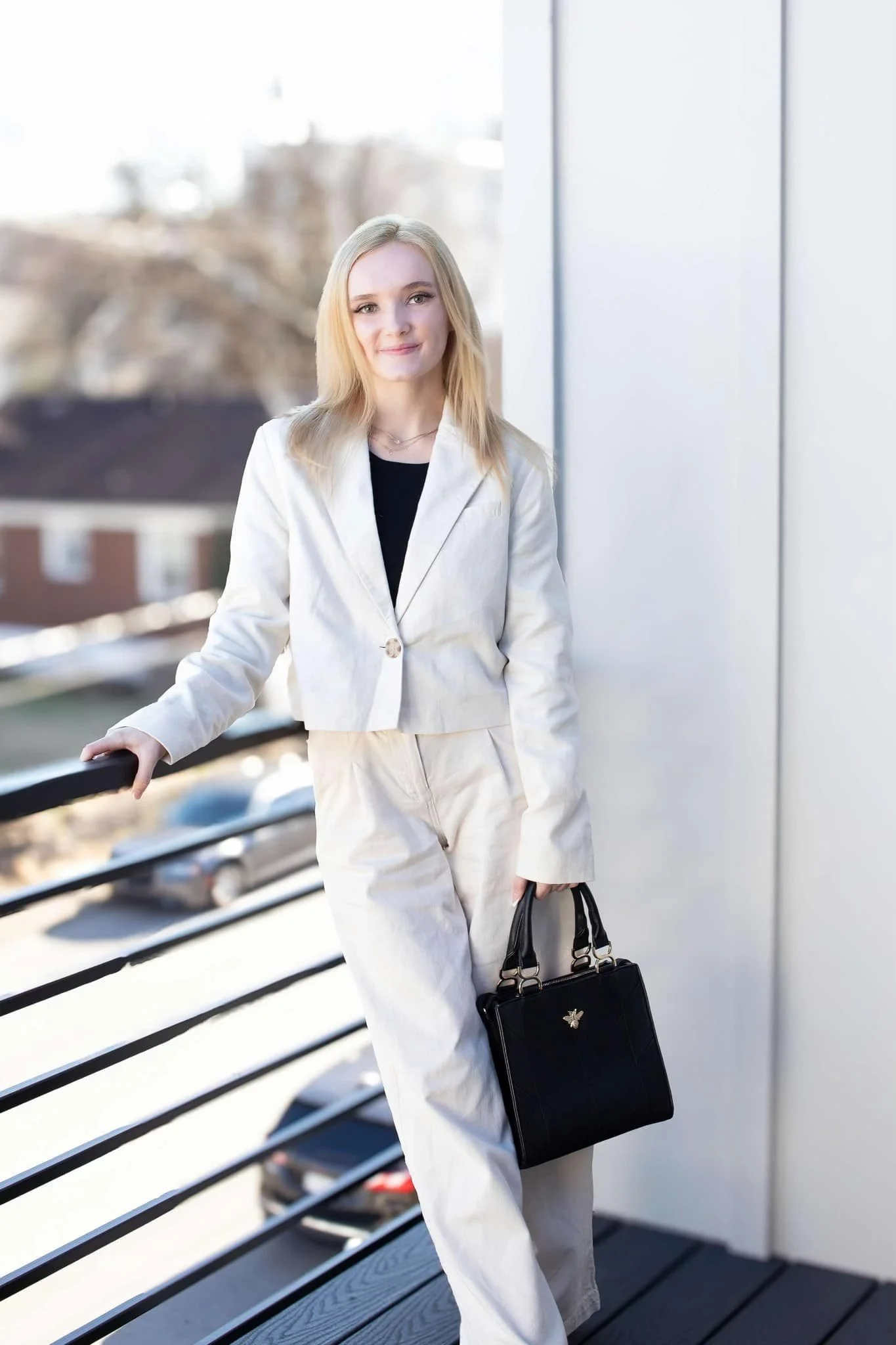 A young woman with blonde hair wearing a white blazer and pants, holding a black handbag, standing outdoors near a railing on a sunny day.