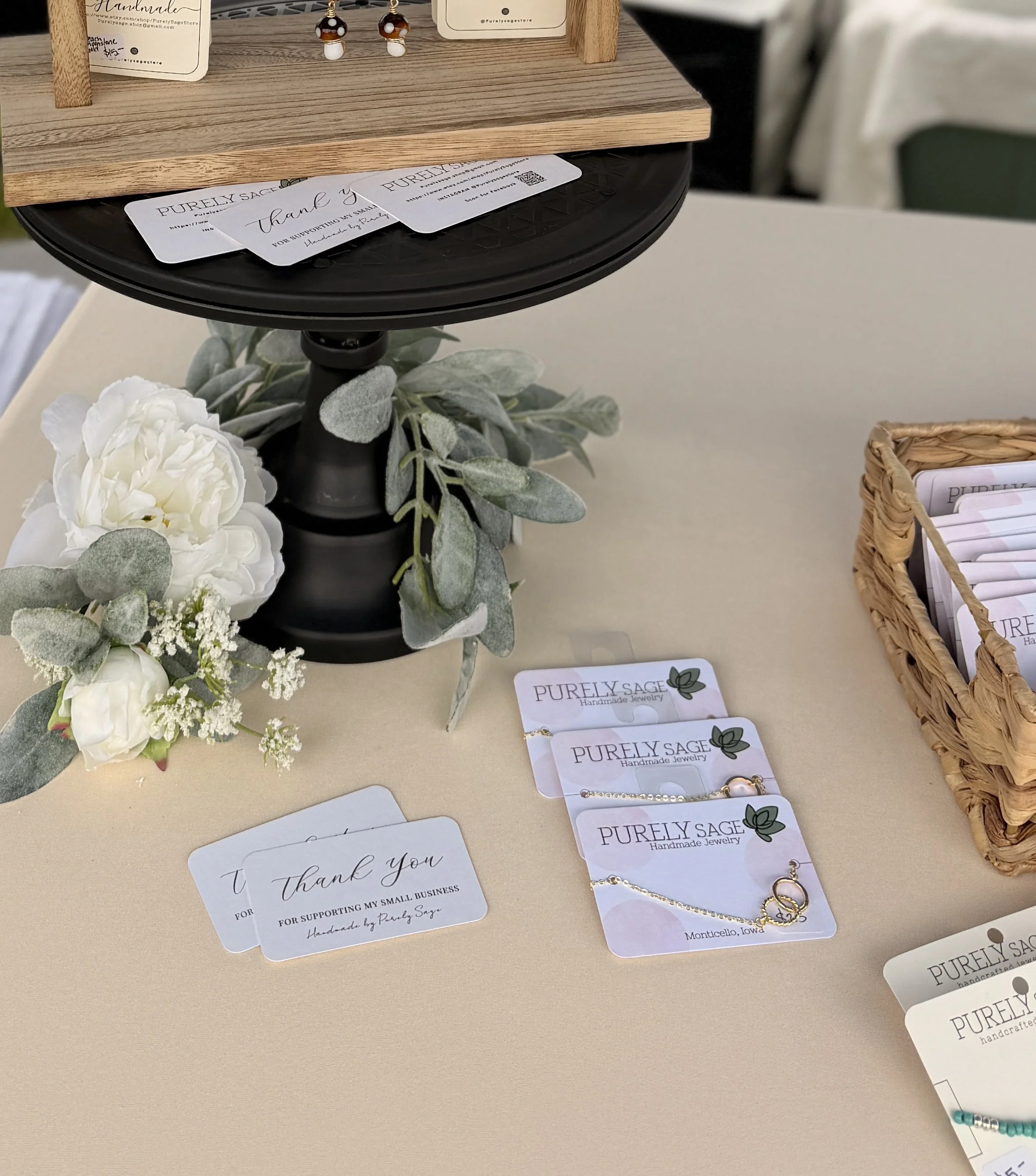 A display of handmade jewelry from Purely Sage, including earrings and bracelets on a table decorated with gray-green foliage and white flowers, with a thank you note and business cards visible.