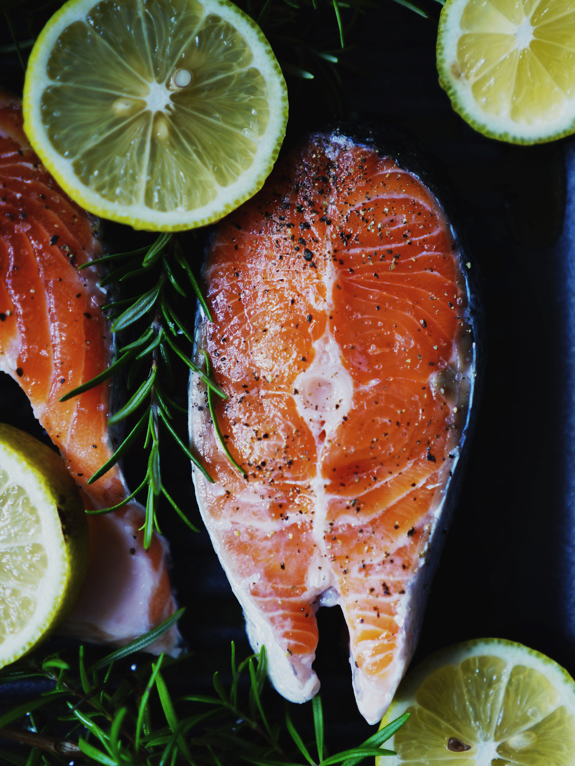 Close-up of raw salmon fillet sprinkled with black pepper, surrounded by lemon slices and rosemary sprigs.