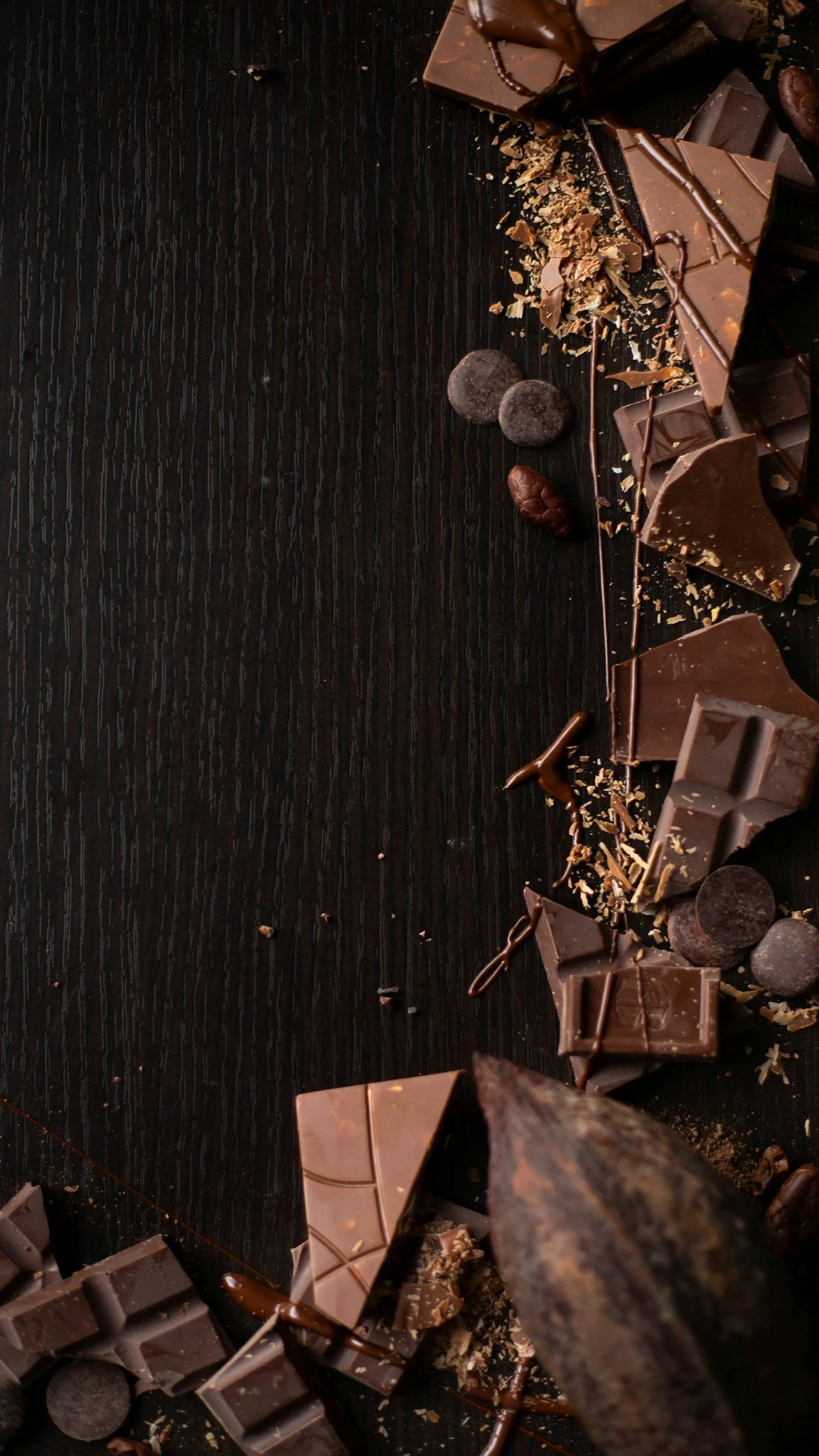 A variety of broken chocolate pieces, chocolate chips, chocolate shavings, and chocolate drizzles scattered on a dark wooden surface.