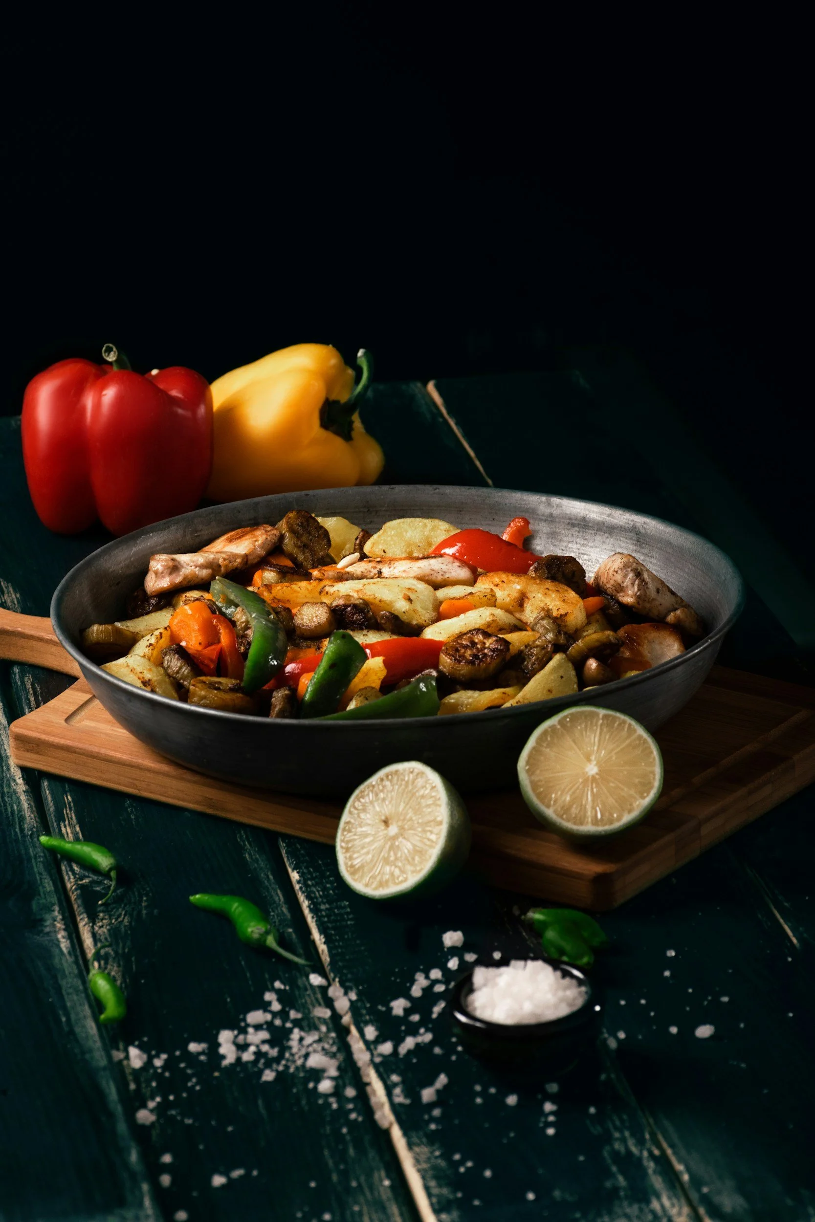 A bowl of roasted vegetables including potatoes, bell peppers, and mushrooms on a wooden board, with fresh lemon halves, green chili peppers, and coarse salt nearby on a dark wooden surface. In the background, there are whole red and yellow bell peppers and a blackboard.
