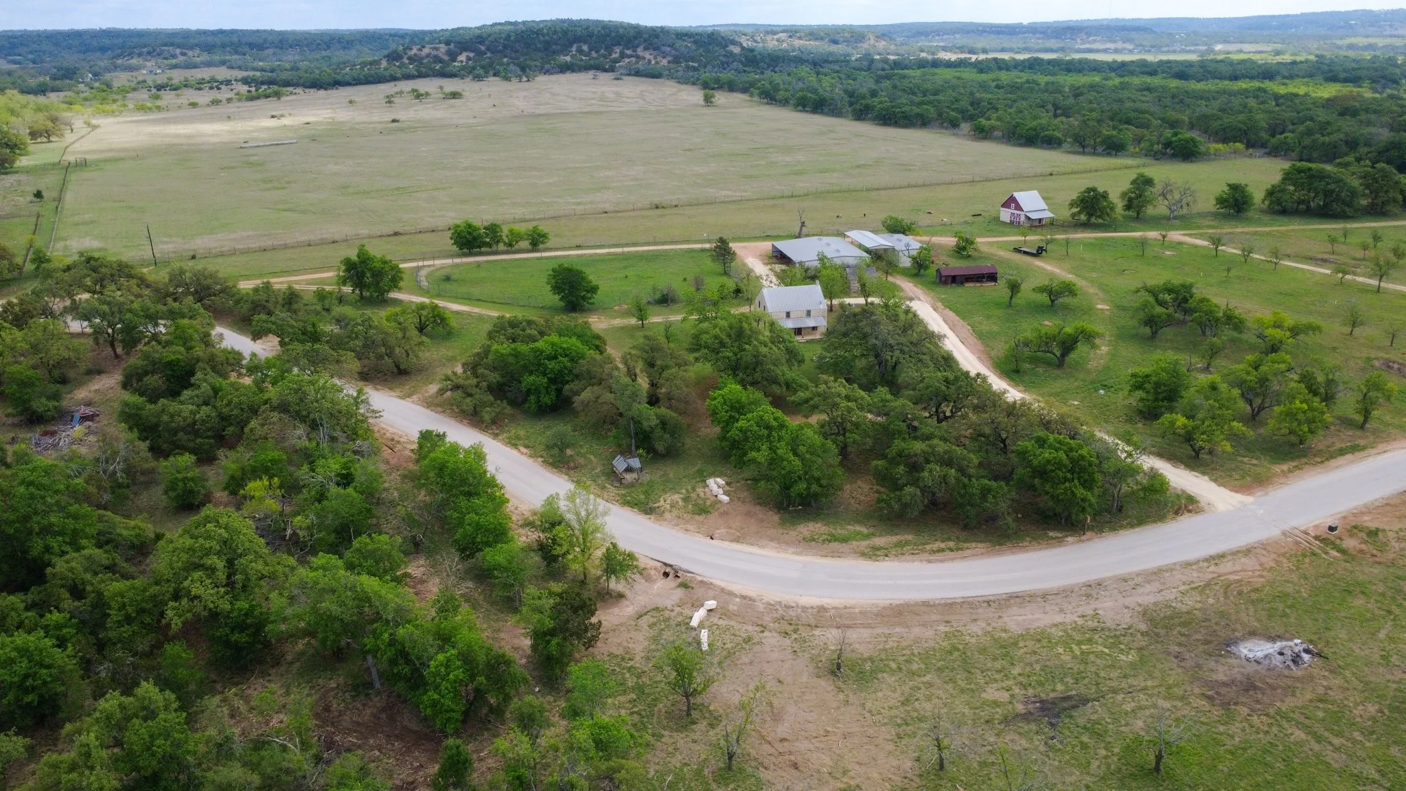 Aerial view of a rural landscape with a farmhouse, other buildings, a winding dirt road, trees, and green fields surrounded by forested hills under a cloudy sky.
