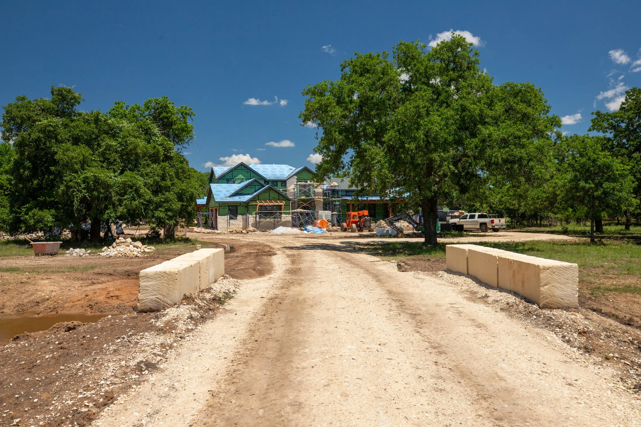 A construction site with a house being built, surrounded by large green trees under a blue sky with some clouds.